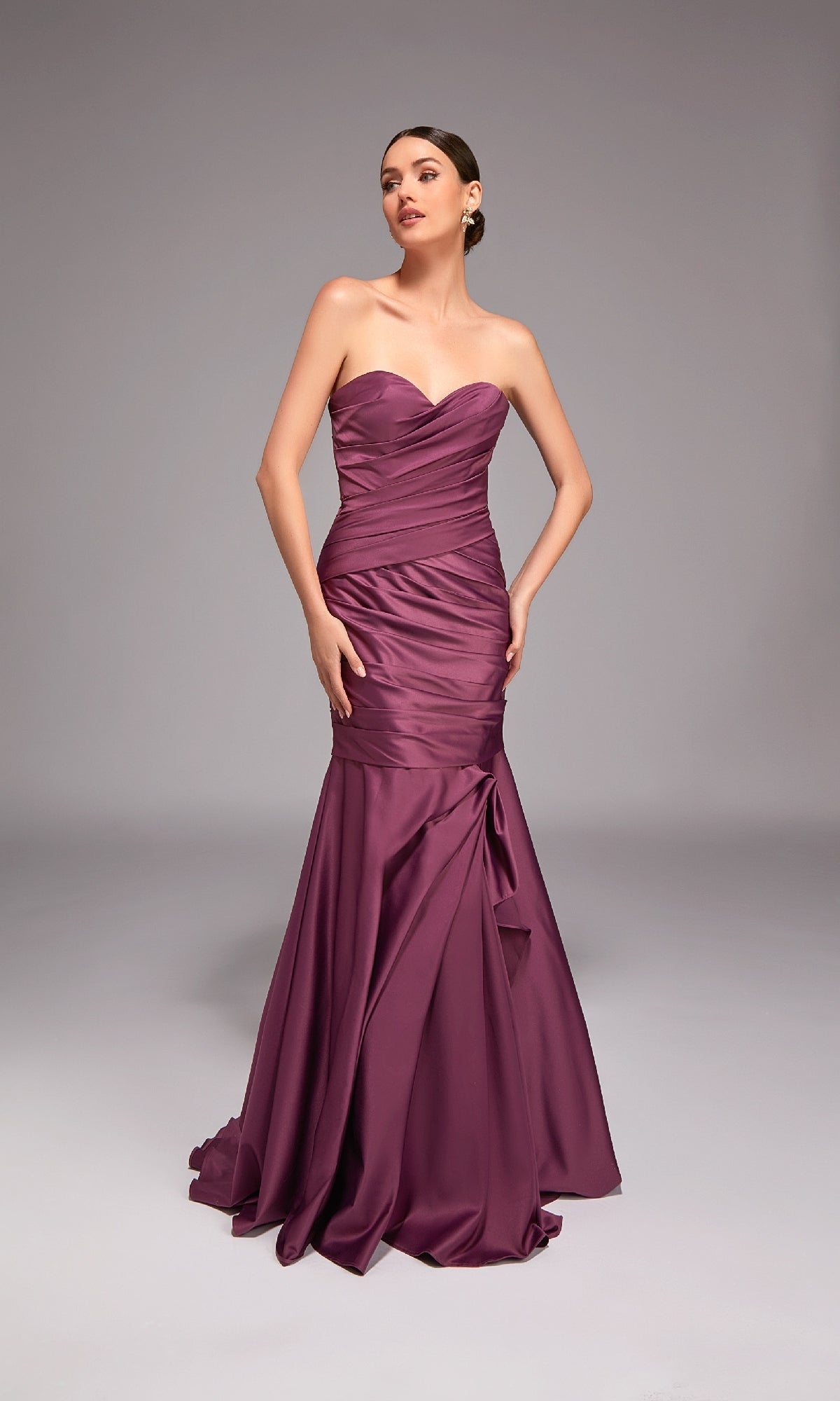 A woman poses against a gray background in the Alyce 27836, a strapless long formal dress with a sweetheart neckline, fitted mermaid skirt, ruched detailing, and slight flare at the hem in purple.