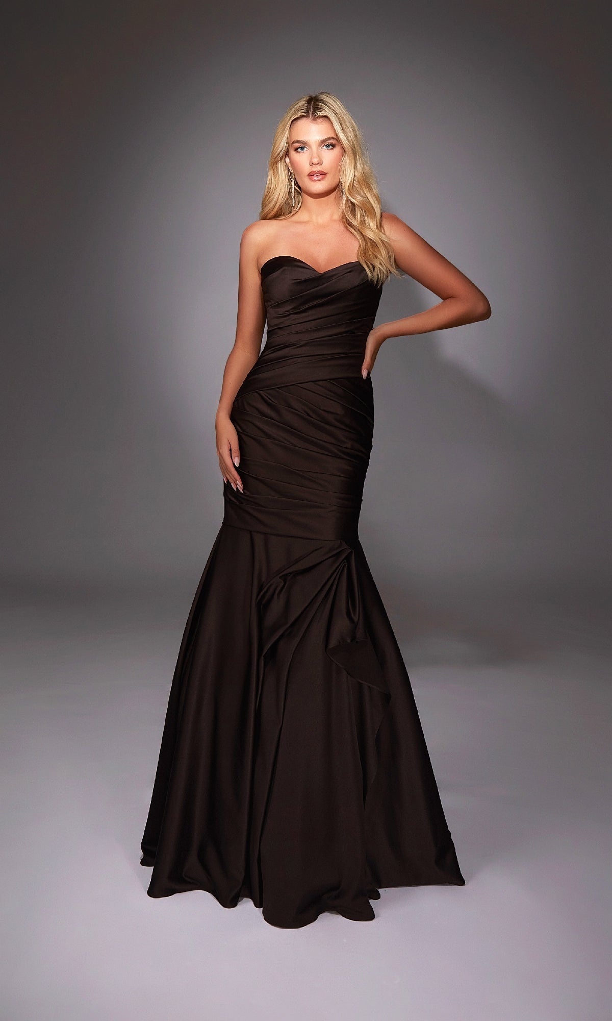 A woman poses in the Alyce 27836 long formal dress, featuring a strapless fitted bodice and dramatic mermaid skirt, the dark brown gown flowing elegantly to the floor against a plain gray backdrop.