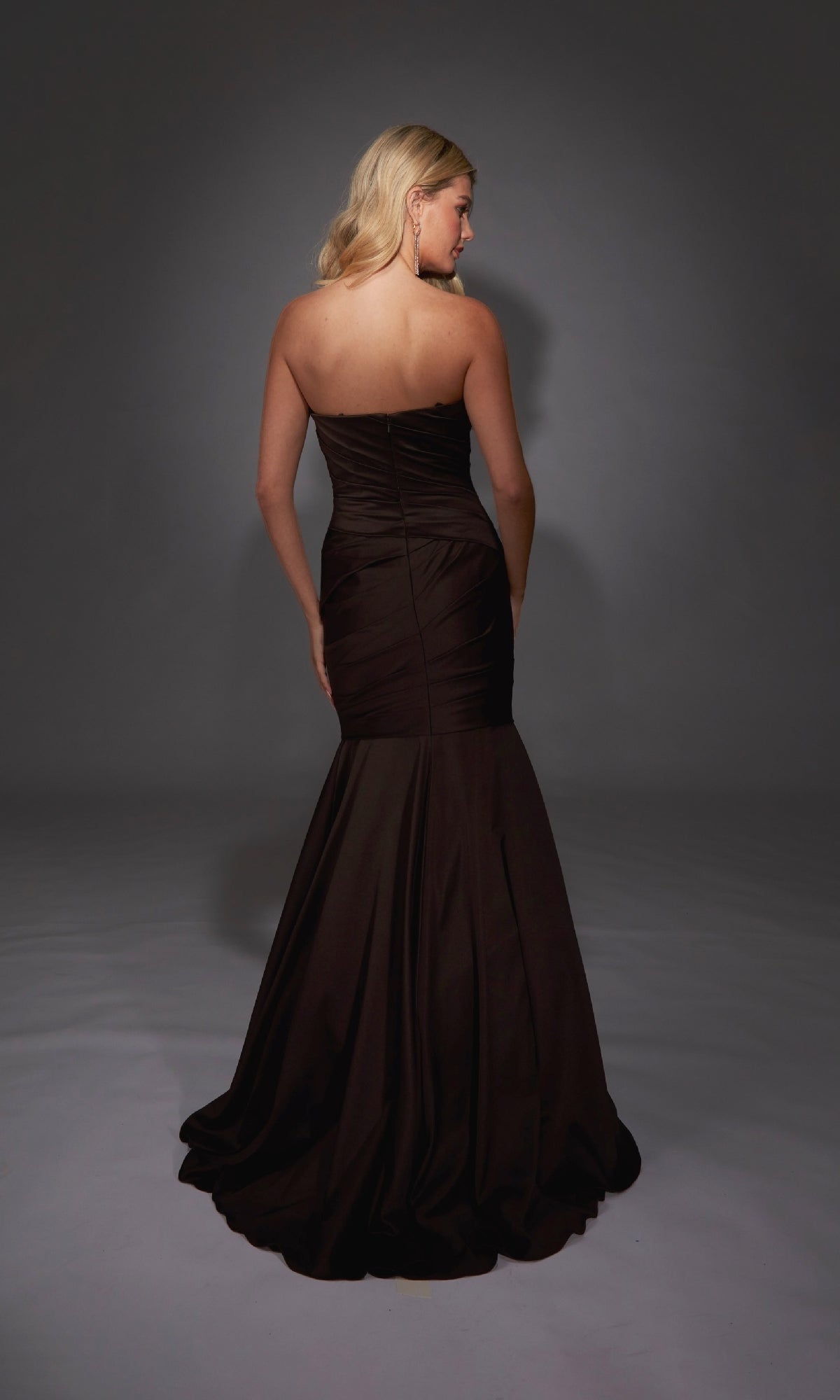 A woman stands facing away in the Alyce 27836 long formal dress—a strapless, floor-length black gown with a sweetheart neckline and fitted mermaid skirt—elegantly flowing in the dimly lit studio.