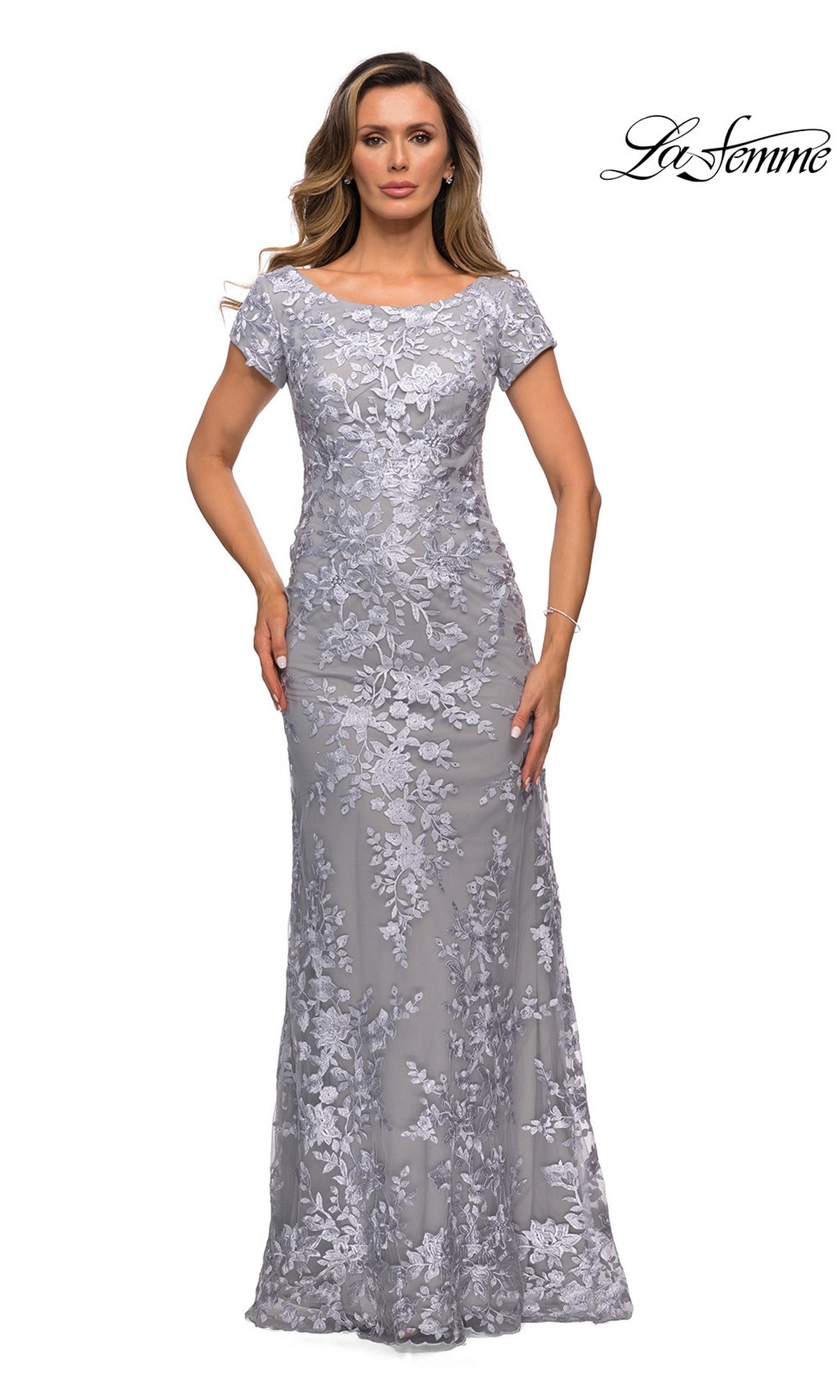 A woman models the Long Formal Dress: La Femme 27842 in silver—a fitted, floor-length sheath with floral lace—posing on white, “La Femme” logo top right.