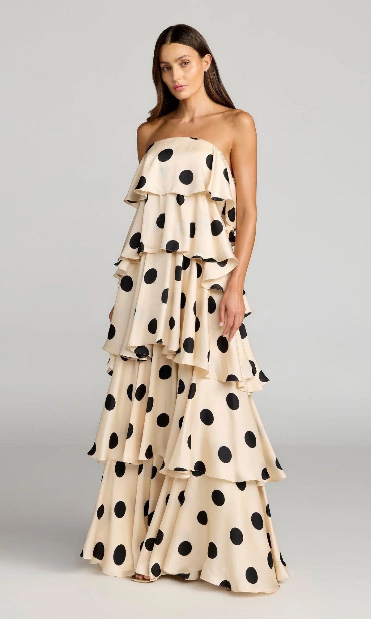 A woman stands against a plain background wearing the Long Formal Dress: Mac Duggal 27845, a floor-length, strapless gown in cream with large black polka dots and tiered ruffles.