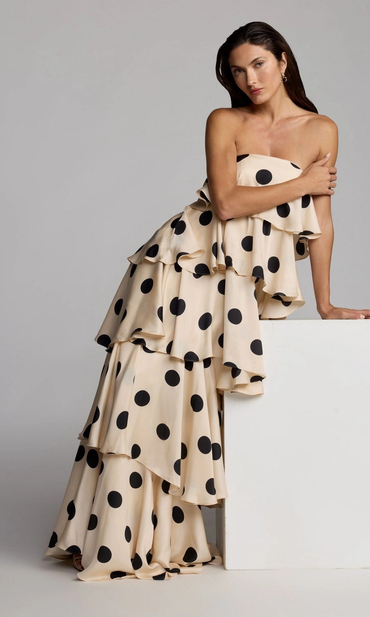 A woman wearing the Long Formal Dress: Mac Duggal 27845—a strapless, tiered cream gown adorned with large black polka dots—leans against a white rectangular block, striking a pose for a studio fashion photo.