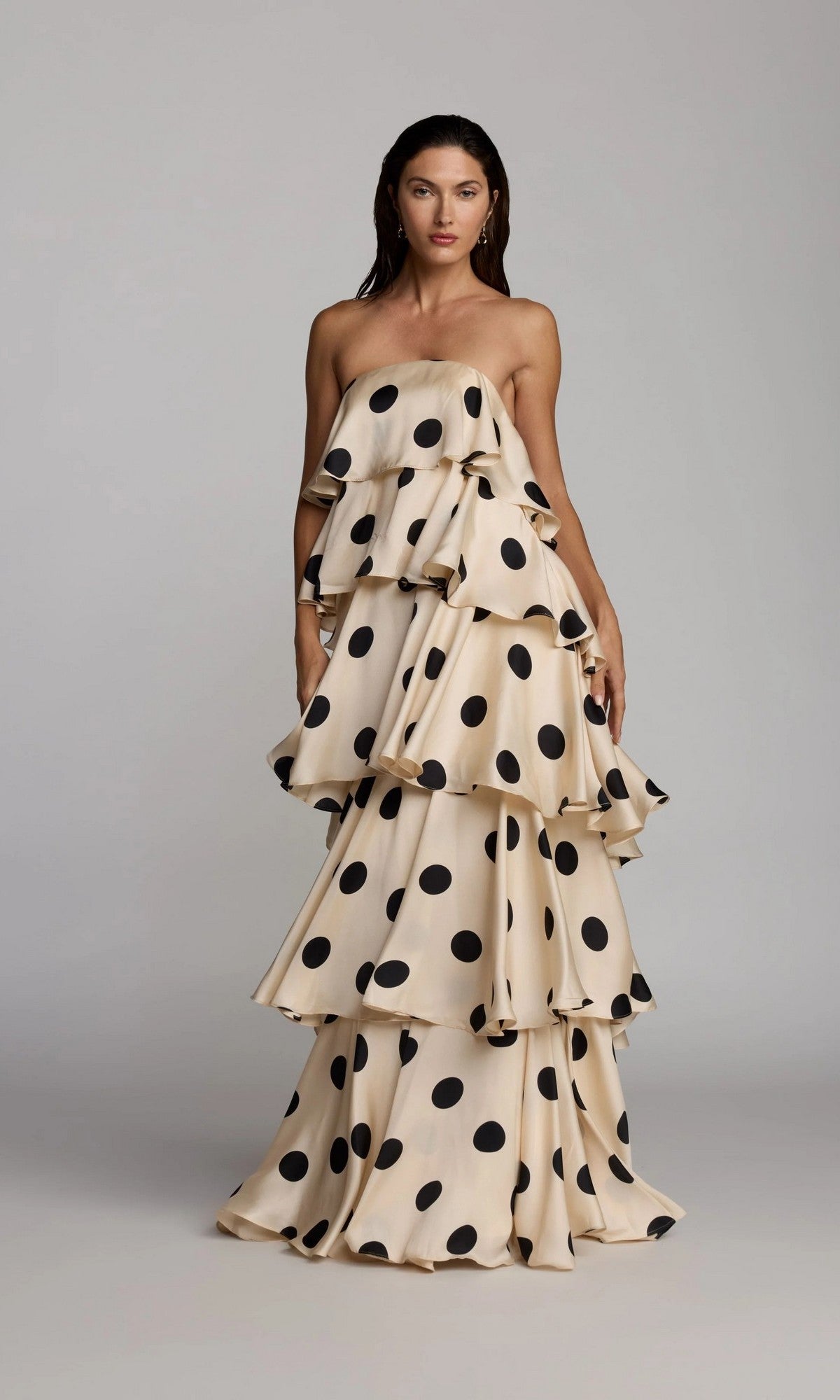 A woman wearing the Mac Duggal 27845 long formal dress, featuring a strapless neckline and tiered ruffle design in cream with large black polka dots, stands against a plain gray background.