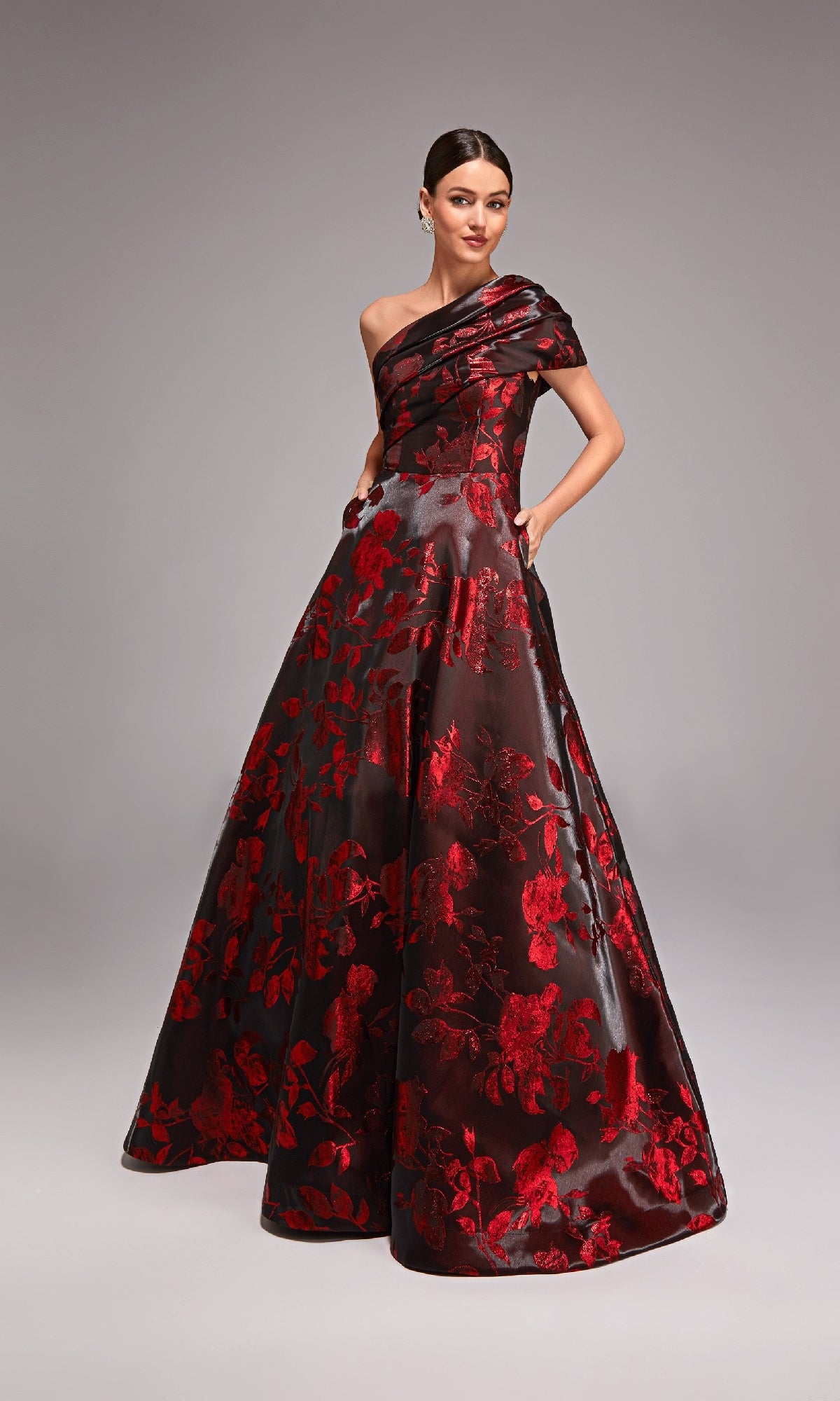 A woman models the Alyce 27858 long formal dress, a floor-length jacquard gown with a black base, red floral pattern, and striking one-shoulder design, set against a plain gray background.