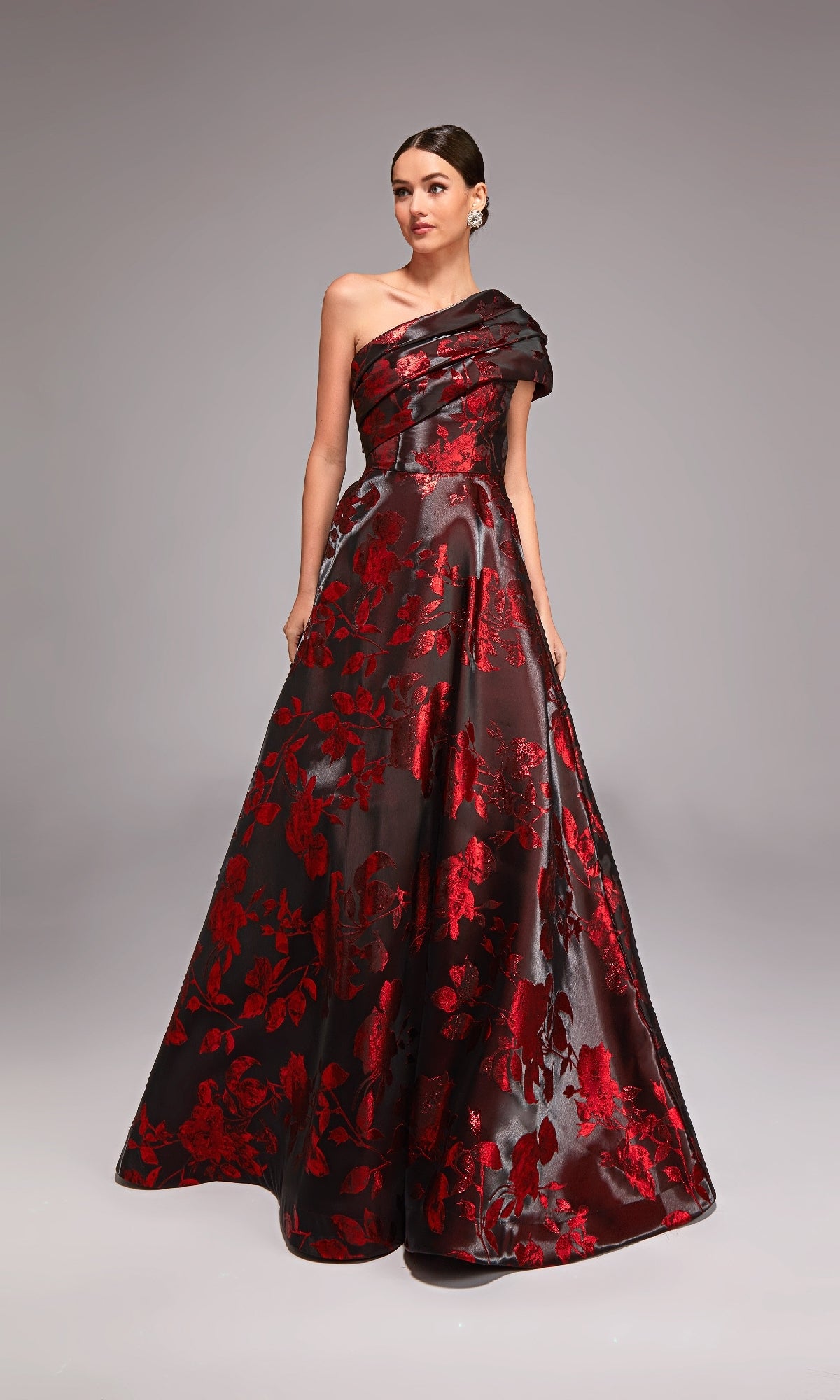 A woman in the Alyce 27858 long formal dress, featuring a one-shoulder jacquard design with red floral patterns, stands against a plain gray background.