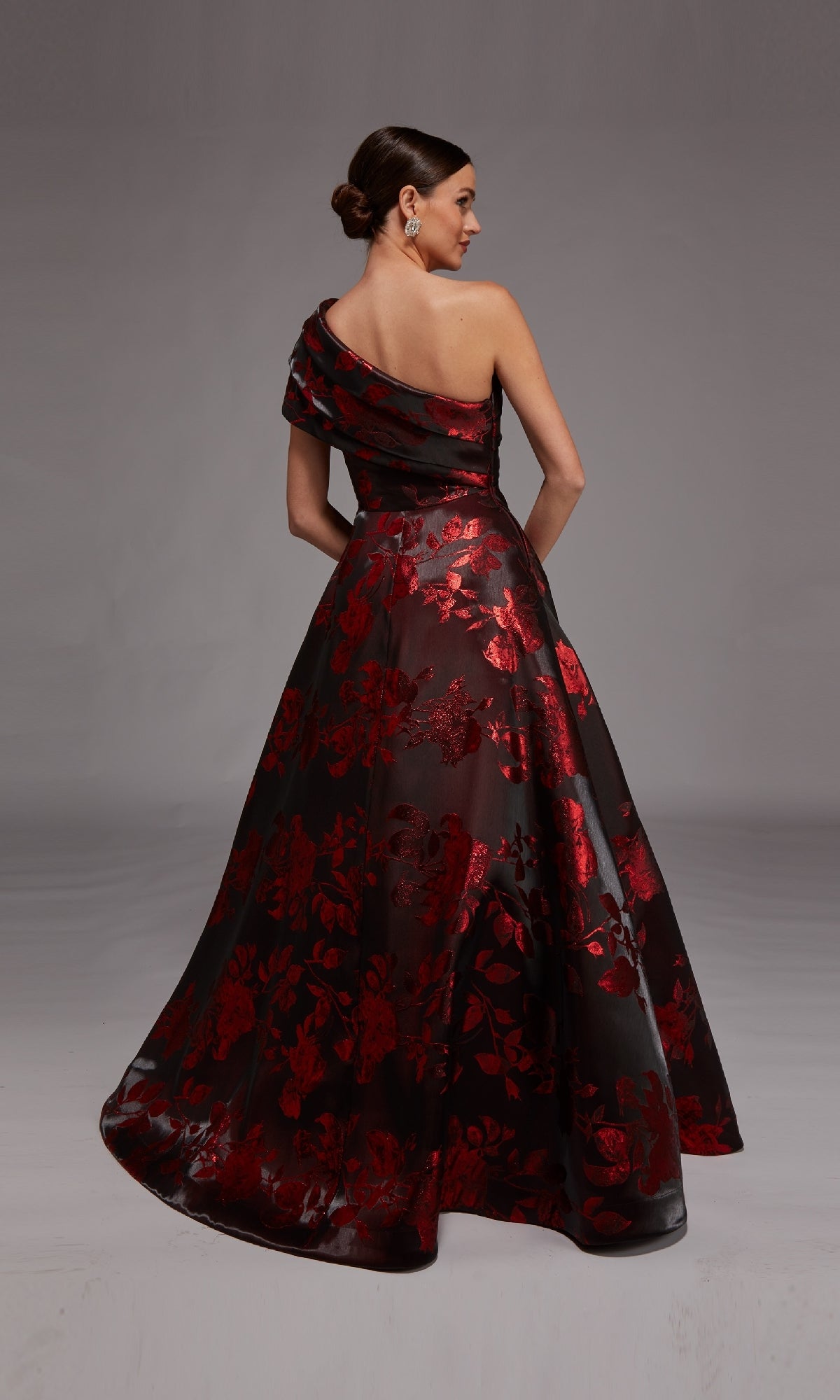 A woman models the Alyce 27858 long formal dress—a floor-length, one-shoulder gown featuring a black jacquard base with red floral patterns—set against a plain gray background.