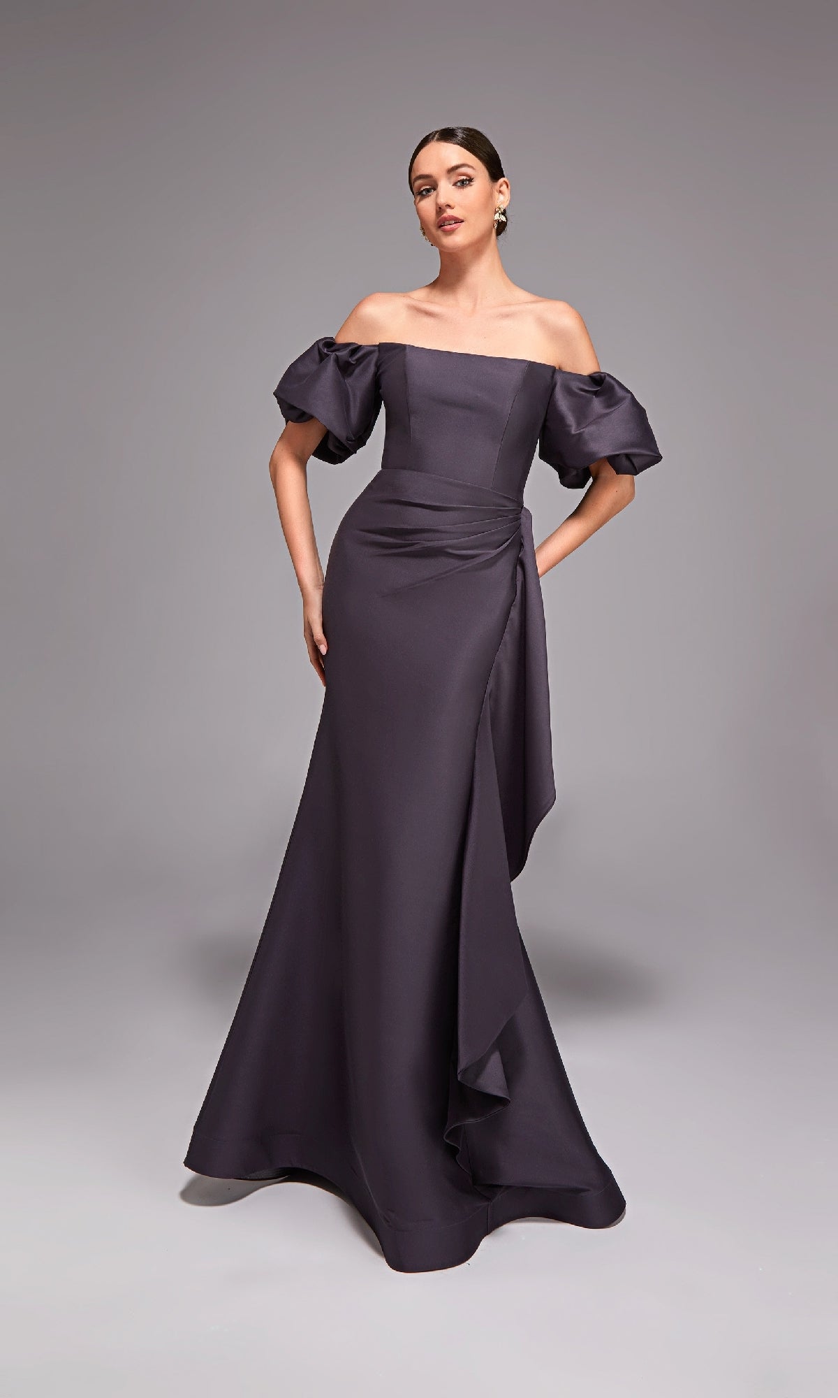 A woman poses in the Alyce 27863 long formal dress—an off-the-shoulder style with puff sleeves, a draped waist, and a fitted silhouette—against a plain gray background.