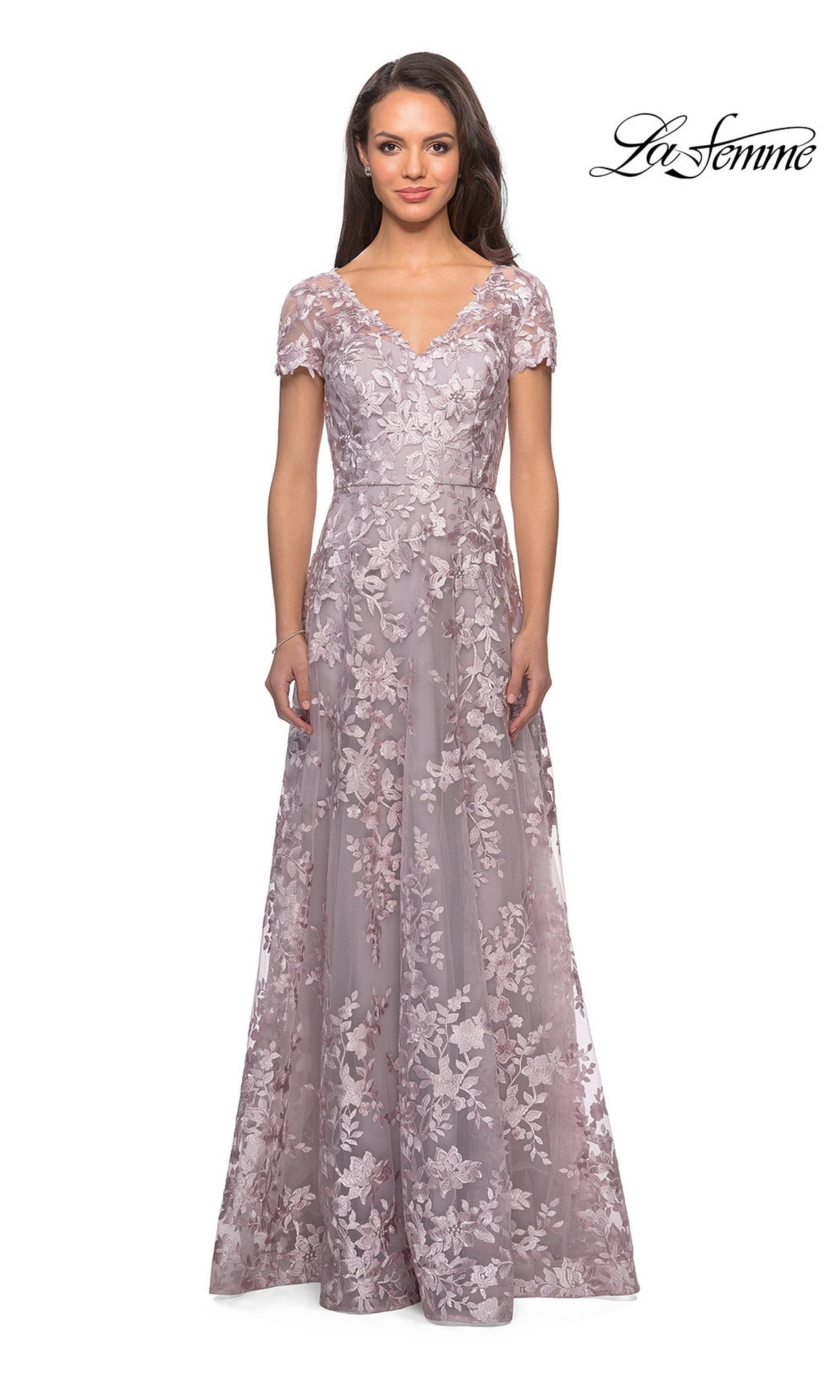 A woman wearing a floor-length, short-sleeve, antique blush La Femme 27870 mother of the bride dress with delicate floral lace patterns stands against a white background. This modest formal gown beautifully showcases intricate detailing, while the La Femme logo appears in the top right corner.