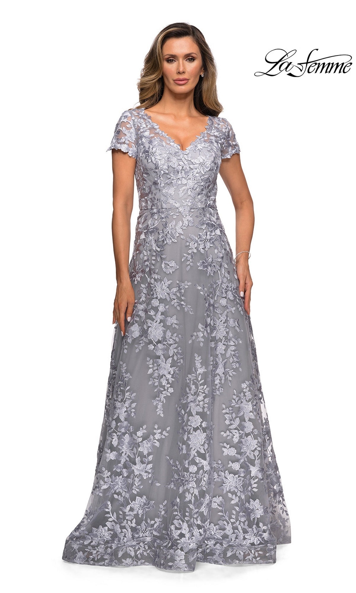 A woman models the La Femme 27870, a long formal dress in platinum featuring short sleeves and floral lace embroidery for an elegant A-line silhouette. The La Femme logo is displayed in the top right corner.