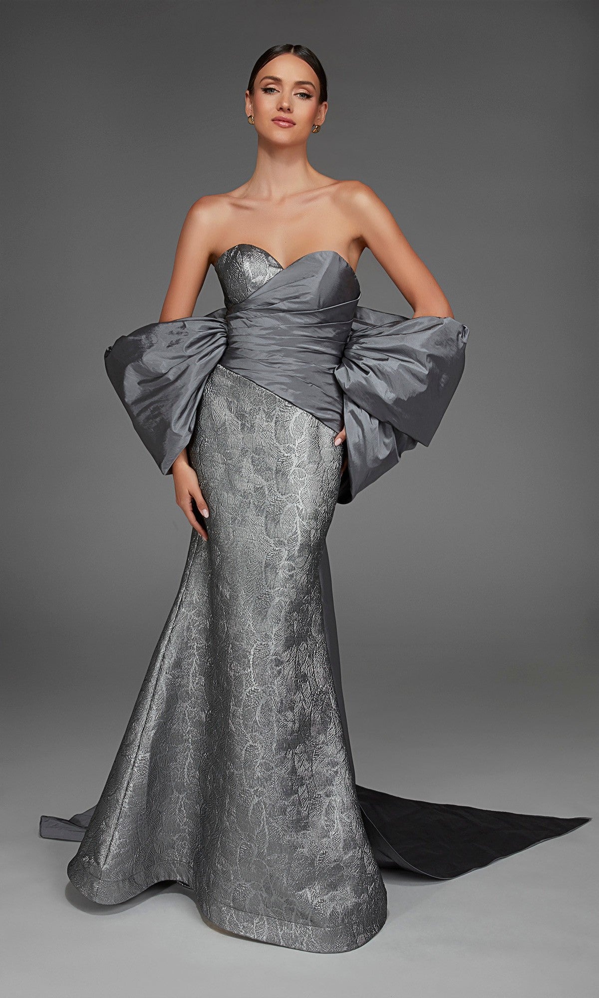 A woman wearing the Alyce 27881 Long Formal Dress in Dripping Diamonds (Silver)—a strapless gown crafted from textured, metallic jacquard fabric with a fitted bodice and dramatic oversized sleeves—stands against a plain gray background.
