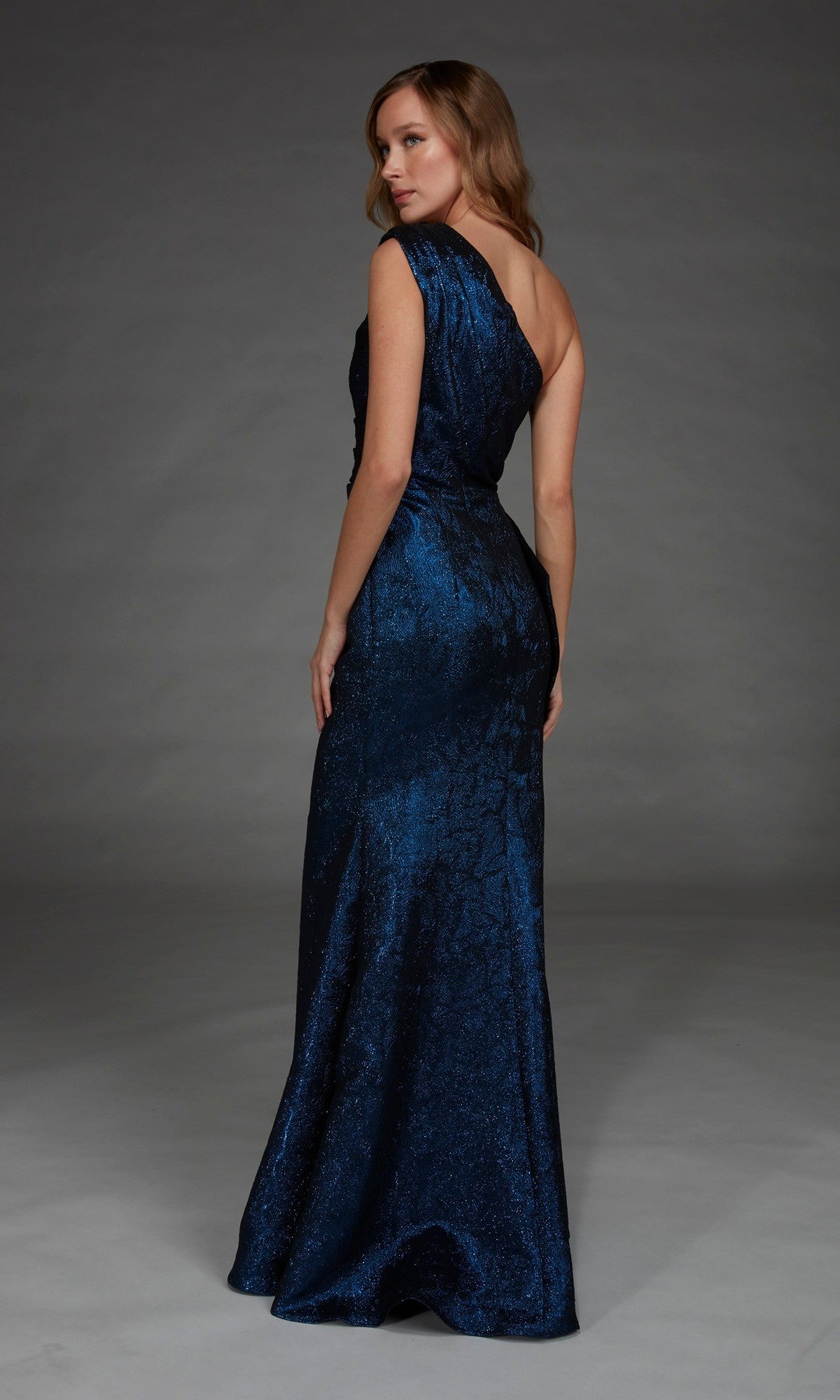 Wearing the Alyce 27885 Long Formal Dress in dark blue, a woman stands with her back slightly turned, gazing over her shoulder.
