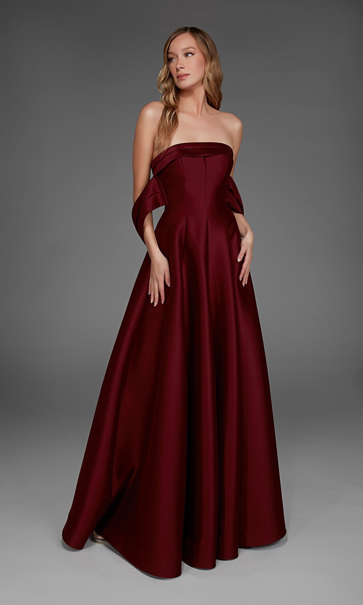 A woman wearing the Alyce 27887 long formal dress in burgundy, featuring a strapless, floor-length design with a fitted bodice and draped fabric at the arms, stands against a plain gray background.
