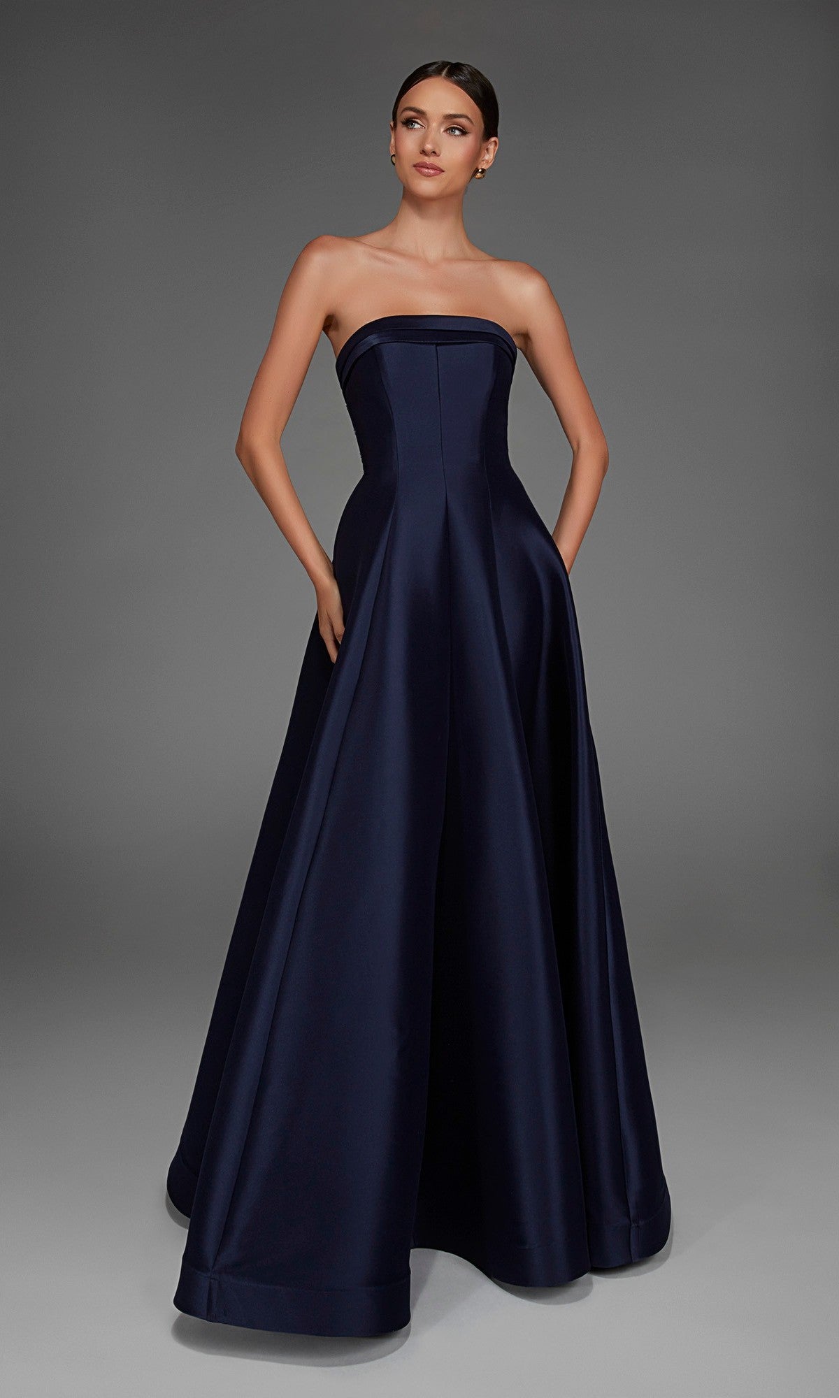 A woman wearing the Alyce 27887 long formal dress—an A-line, strapless, floor-length gown in dark navy with pockets—stands against a plain grey background.