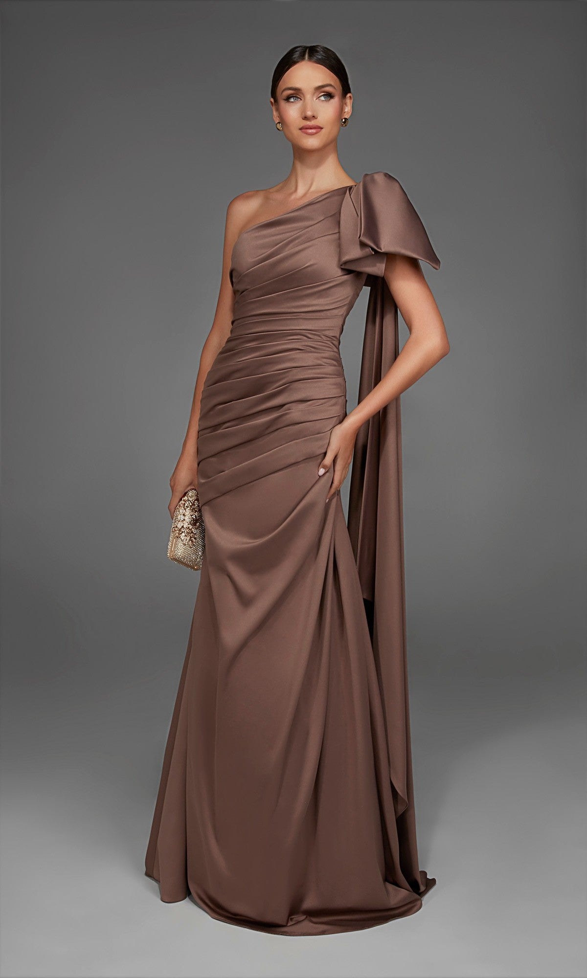 A woman stands against a plain gray background wearing the Alyce 27891 long formal dress in Shadow Grey, a floor-length, one-shoulder satin evening gown featuring a dramatic bow detail. She holds a small gold clutch, making an elegant statement in this figure-flattering formal look.