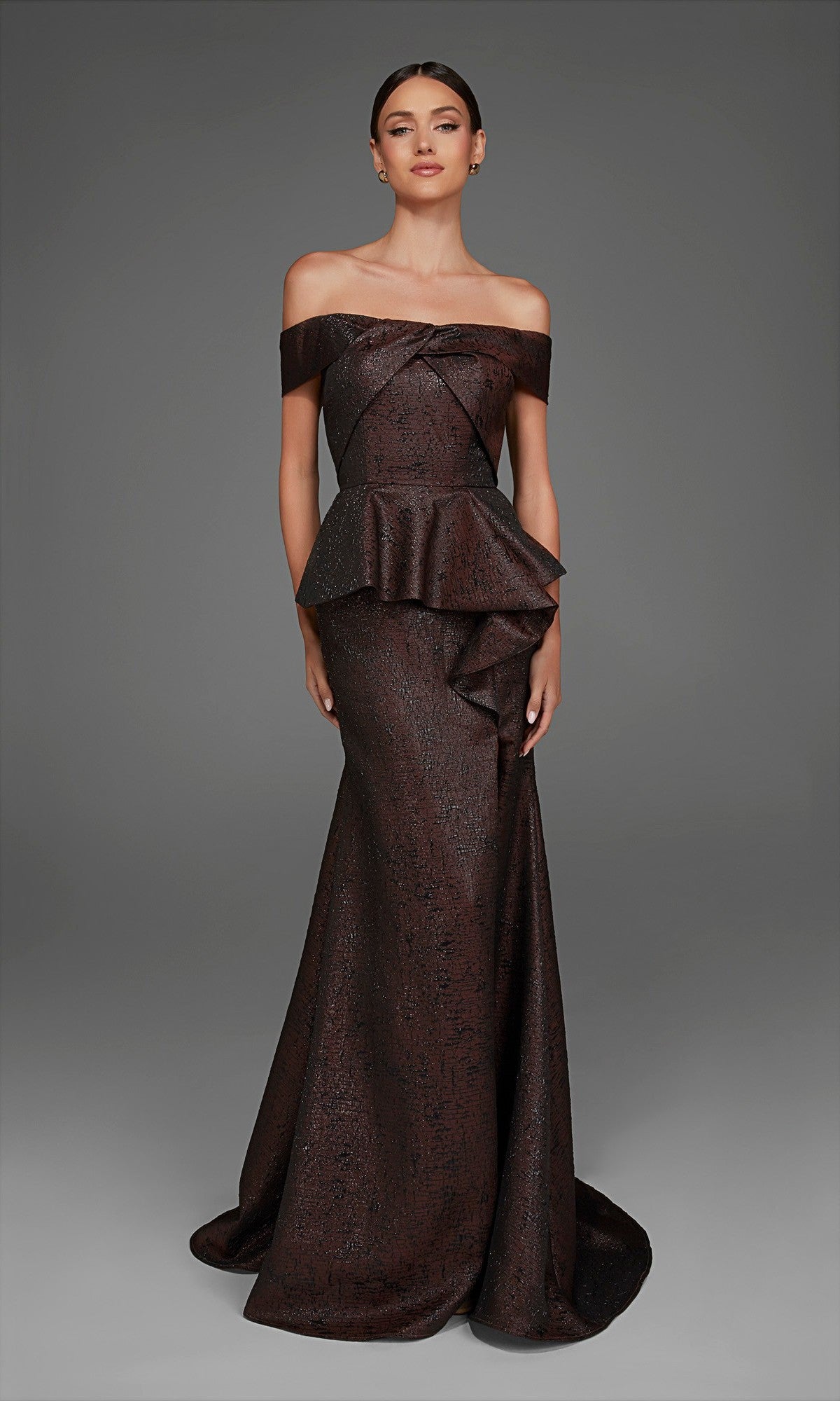 An elegant woman stands gracefully against a plain gray background, wearing the Alyce 27892 Long Formal Dress in espresso—a floor-length gown with an off-the-shoulder neckline, fitted bodice with peplum detailing, and a sophisticated mermaid skirt.