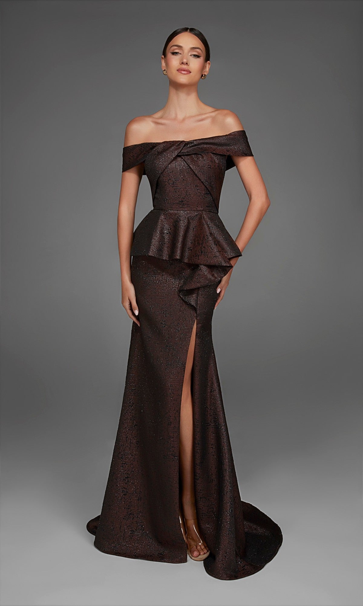 Wearing the Alyce 27892 Long Formal Dress, a woman poses against a plain gray background in a dark off-the-shoulder evening gown, featuring a striking peplum silhouette and mermaid skirt with a thigh-high slit.