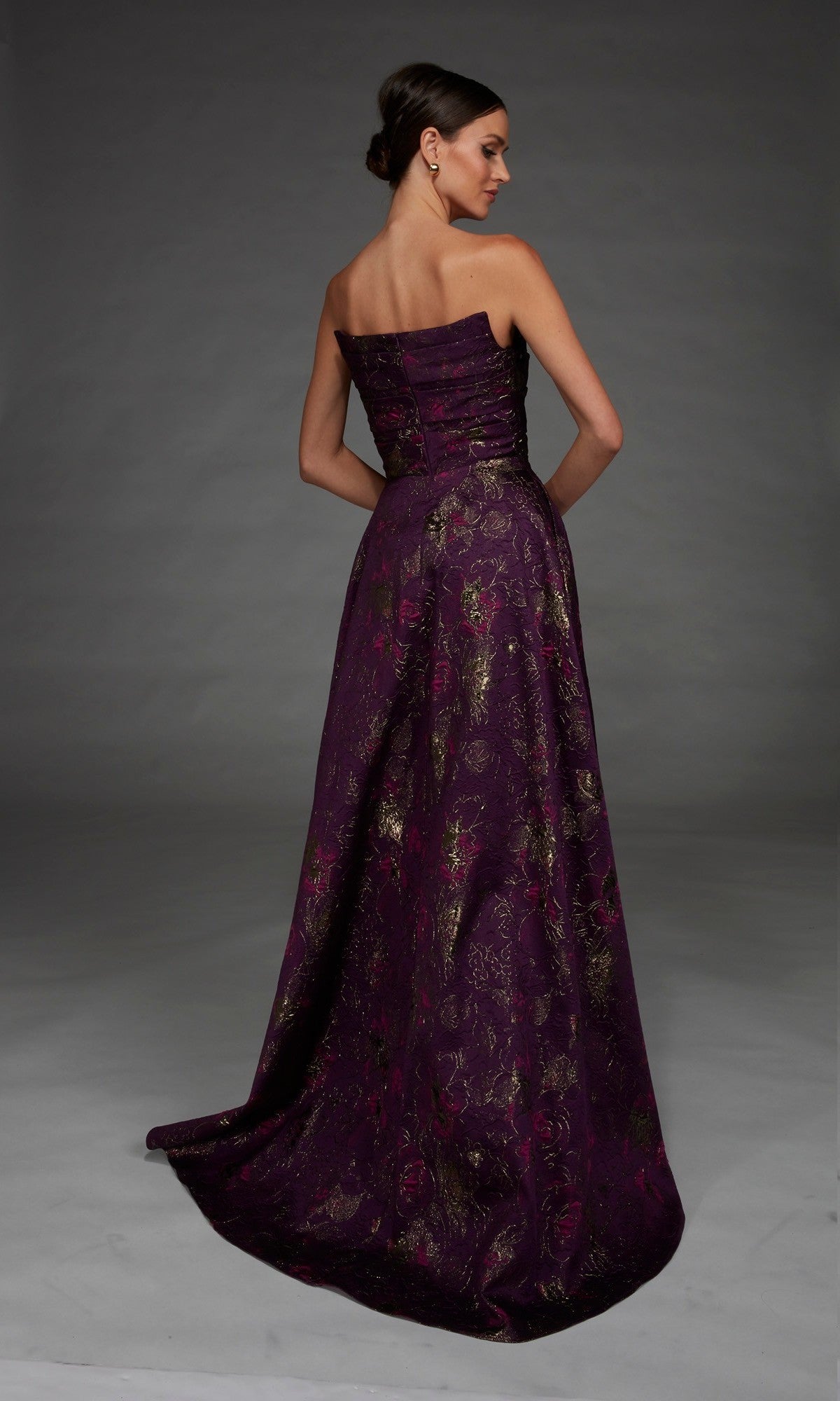 Wearing the Alyce 27893 long formal dress, a woman stands against a gray background, facing away from the camera. The strapless, floor-length gown features a purple floral jacquard fabric adorned with gold and pink patterns.