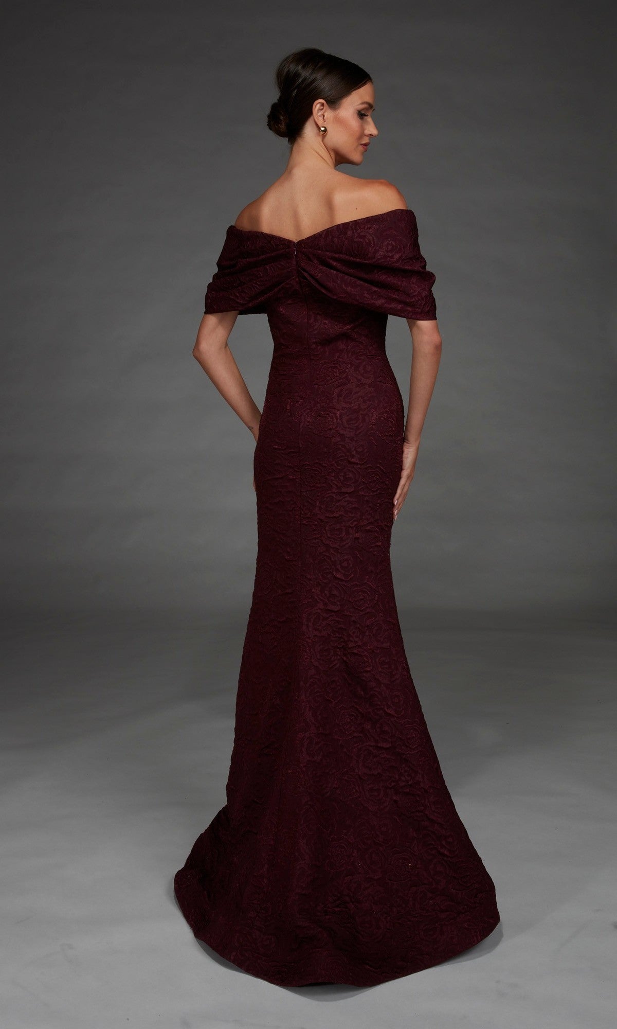 A woman stands with her back to the camera, modeling the Alyce 27894 long formal dress—an off-the-shoulder, floor-length maroon gown featuring a rose-patterned brocade and an elegant mermaid silhouette, set against a plain gray background.