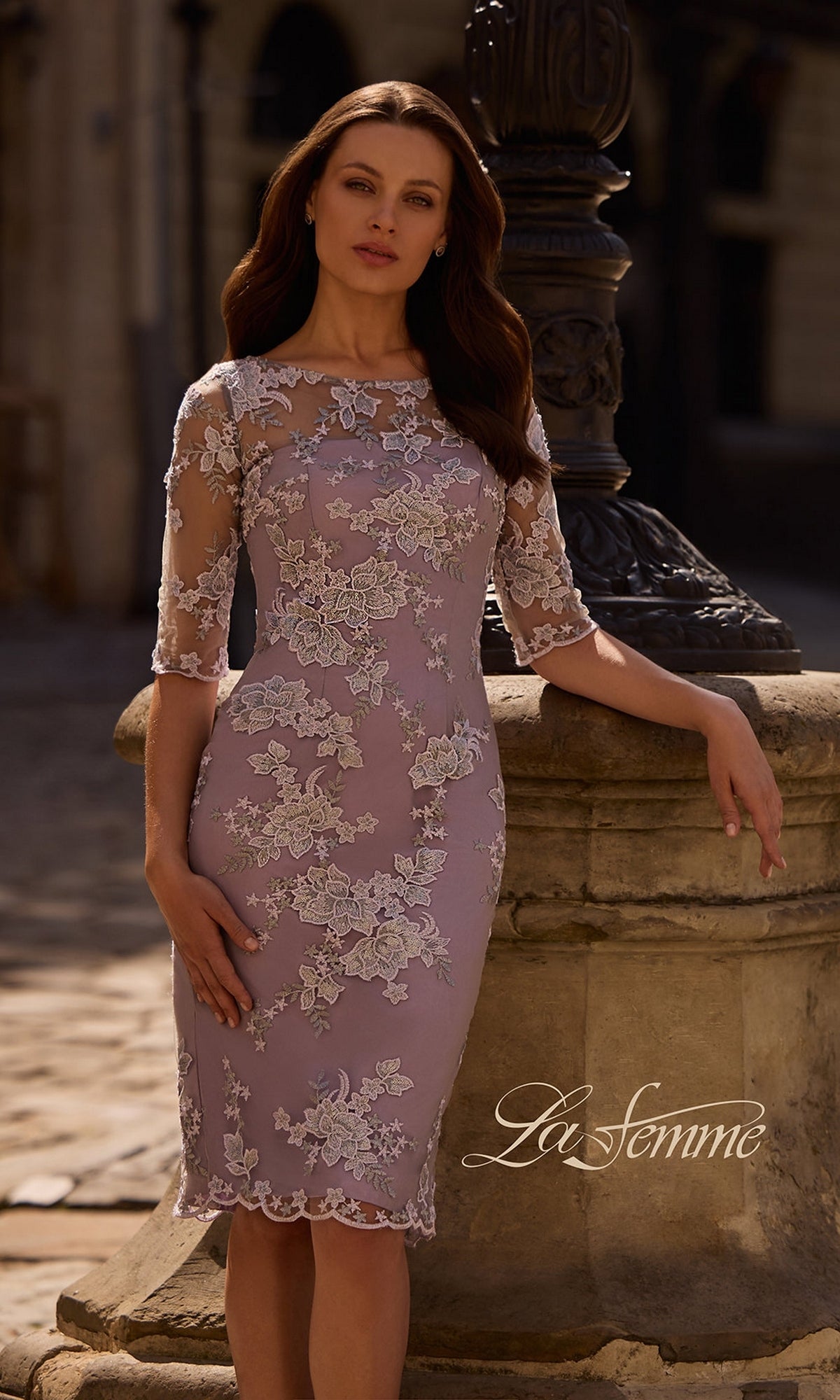 Woman in the Knee Length Wedding Guest Dress: La Femme 27895—lilac floral lace, sheath fit—stands outdoors by a stone wall; “La Femme” logo at bottom right.