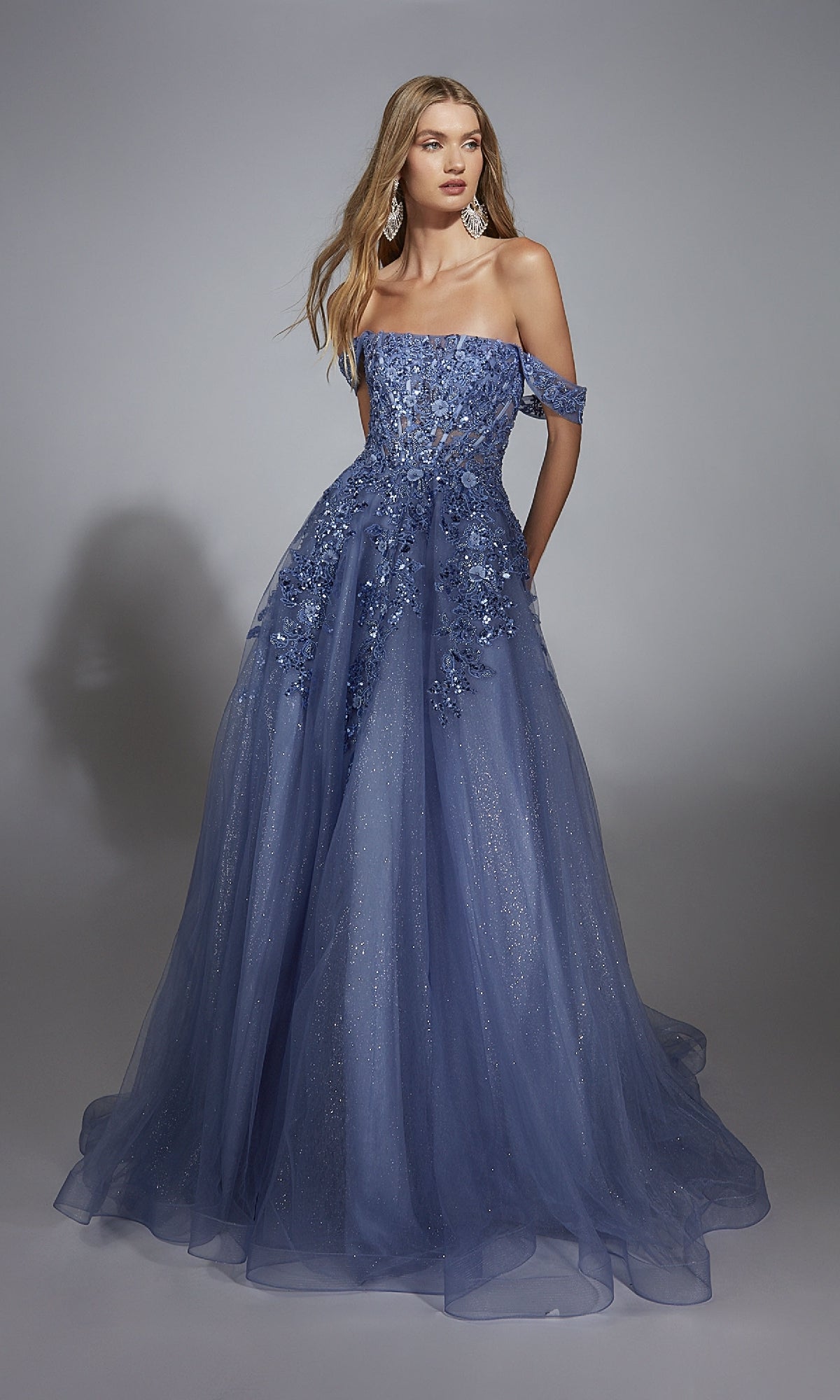 A woman wears the Aerie Couture 278 long prom dress—a blue off-the-shoulder ball gown featuring floral appliqués and lace embroidery on a glitter tulle skirt, standing against a plain gray background.
