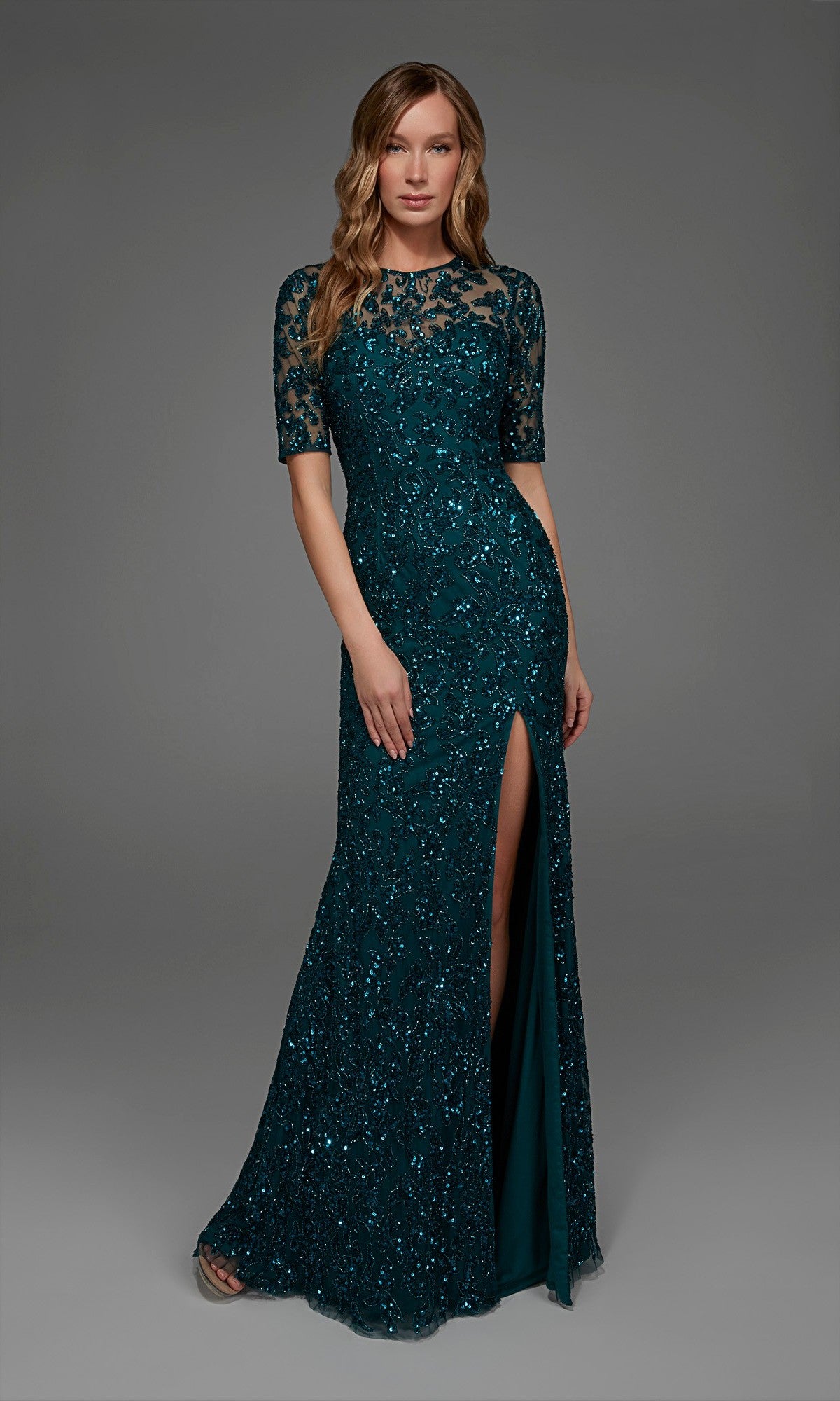 A woman poses against a plain gray background wearing the Alyce 27901 Long Formal Dress in Dragonfly—a teal, floor-length lace gown embellished with beading and sequins, featuring short sleeves, a high slit, and a fitted column silhouette.
