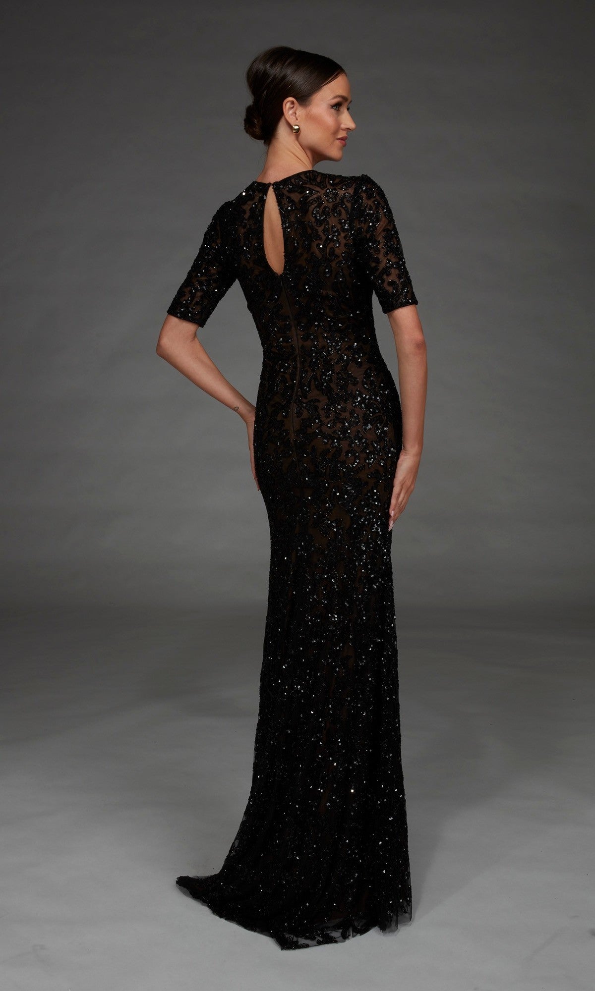 A woman stands with her back to the camera, wearing the Alyce 27901 long formal dress—a floor-length black gown with intricate lace and sequin detailing, short sleeves, and a fitted column silhouette—set against a plain dark background.
