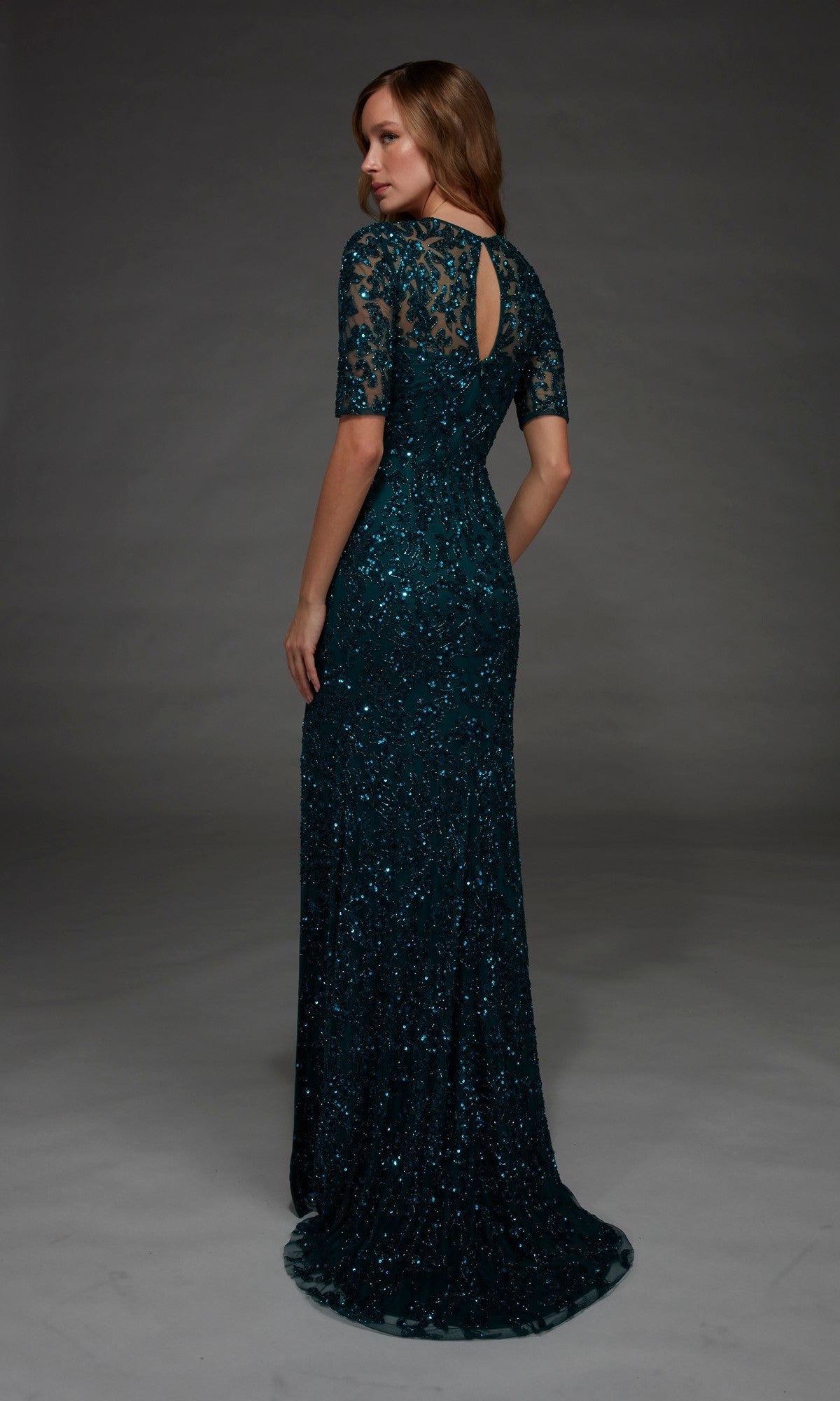 Wearing the Alyce 27901 long formal dress, a woman stands in a dark green, floor-length gown featuring short lace sleeves and a keyhole back. The fitted column silhouette is beautifully embellished with beading and sequins as she faces away, turning her head slightly toward the camera.