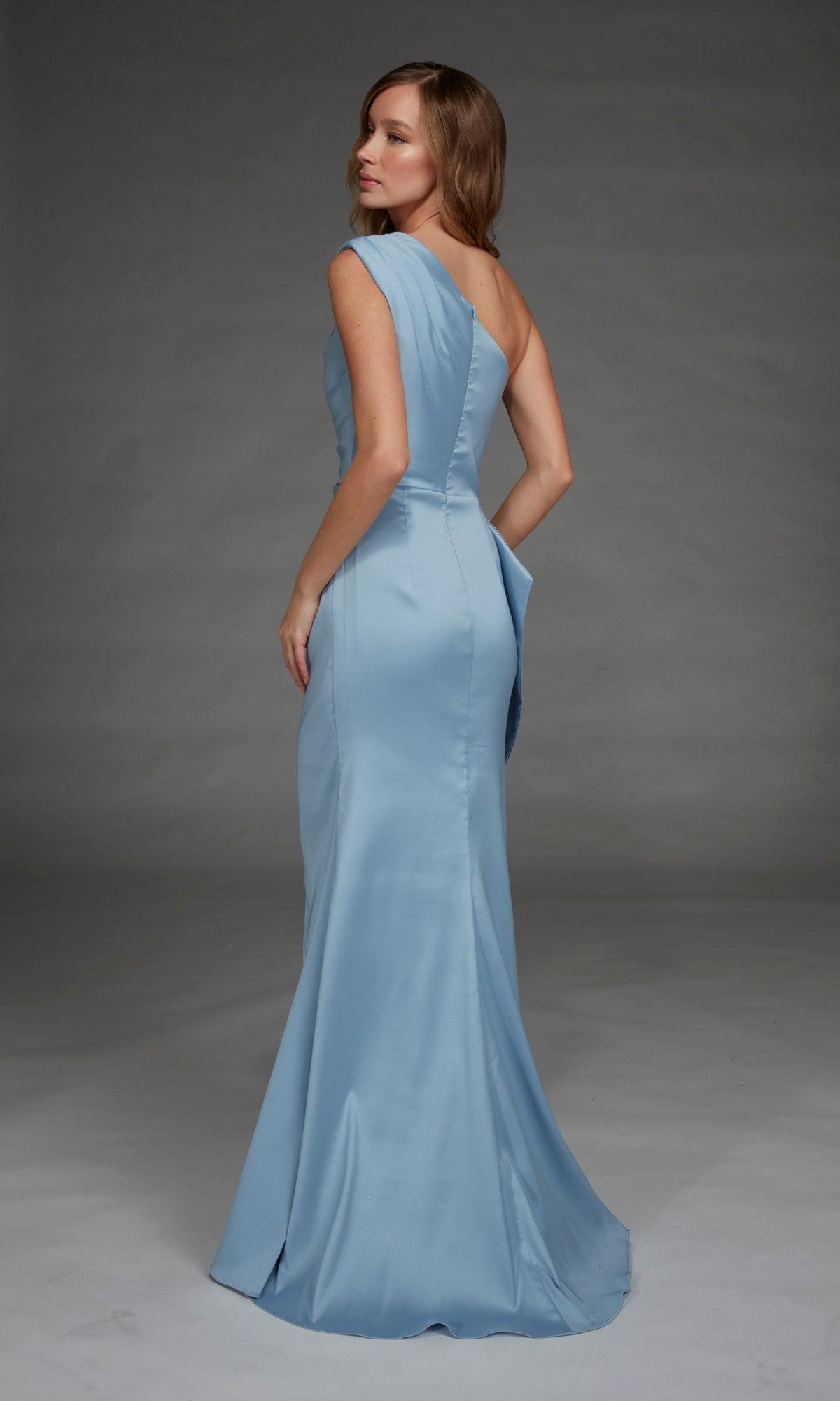 A woman stands against a plain gray background, looking over her shoulder, wearing the Alyce 27913 long formal dress in light blue satin with an elegant one-shoulder neckline.