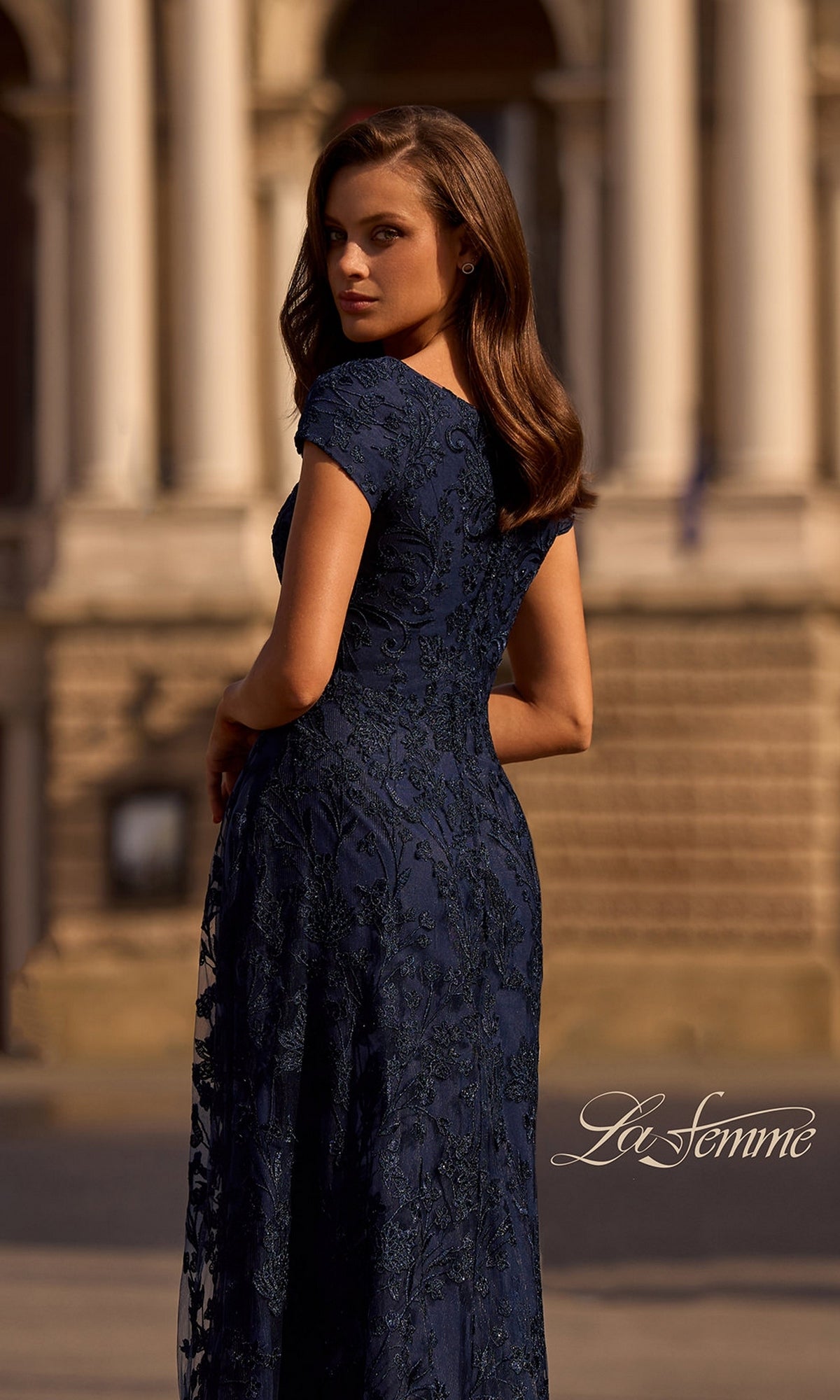A woman wearing the Long Formal Dress: La Femme 27915 poses outside a classical stone building, looking over her shoulder. The La Femme logo is visible in the lower right corner.