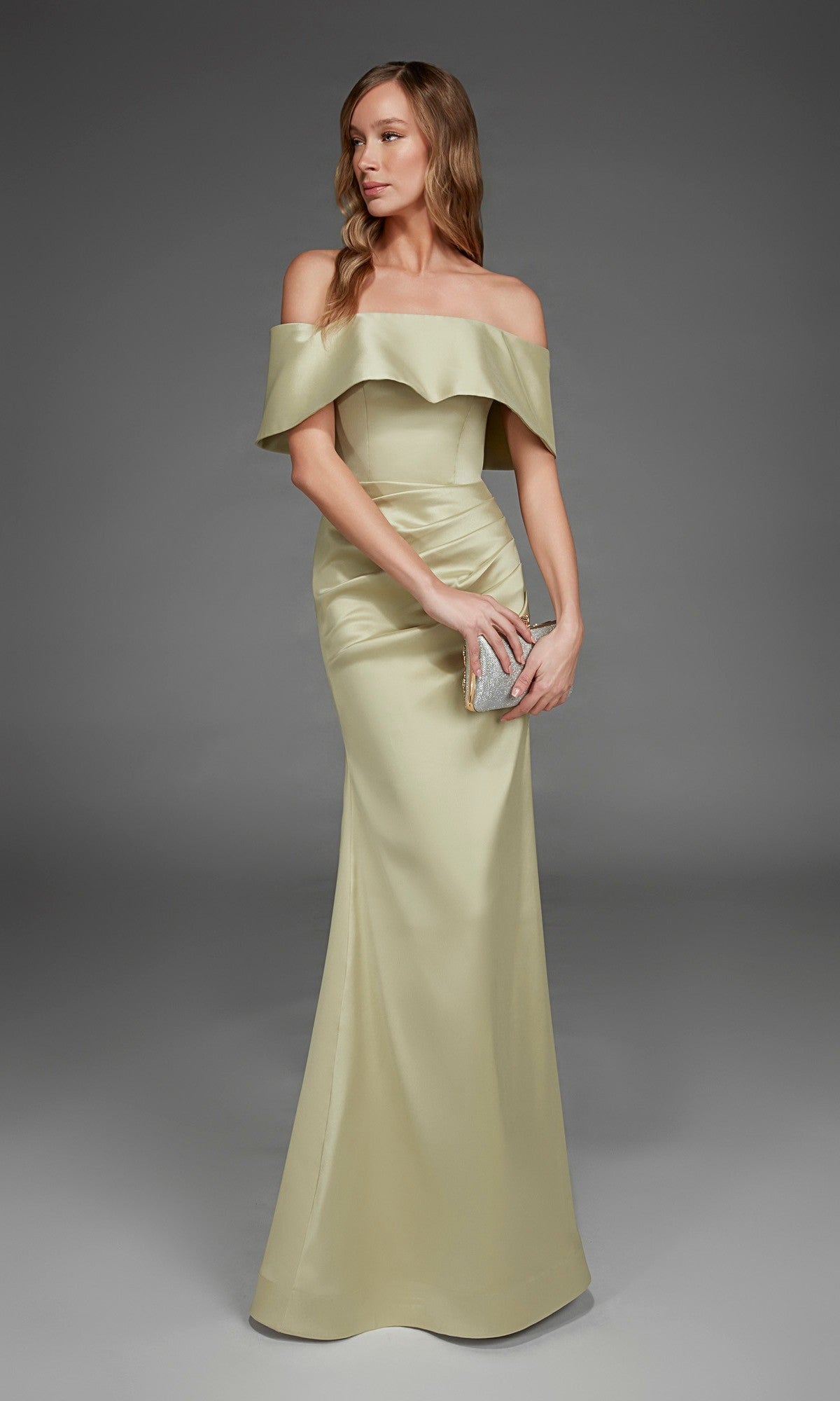 Wearing the Alyce 27916 Long Formal Dress in honeydew, a woman elegantly holds a silver clutch as she stands gracefully against a plain gray background.