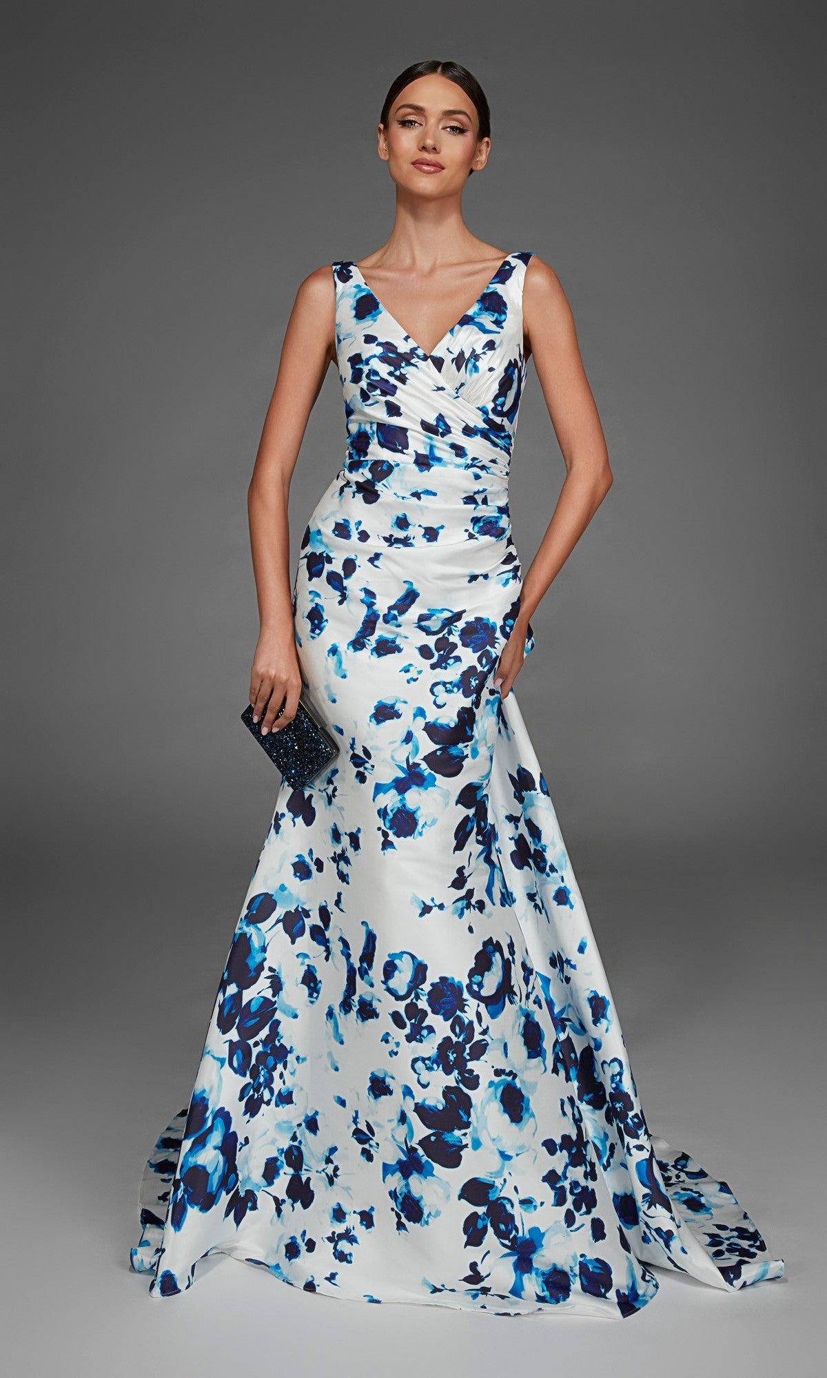 A woman wearing the Alyce 27917 long formal dress in blue/white, a sleeveless floor-length v-neck evening gown with floral patterns, holds a small black clutch and stands against a plain gray background.