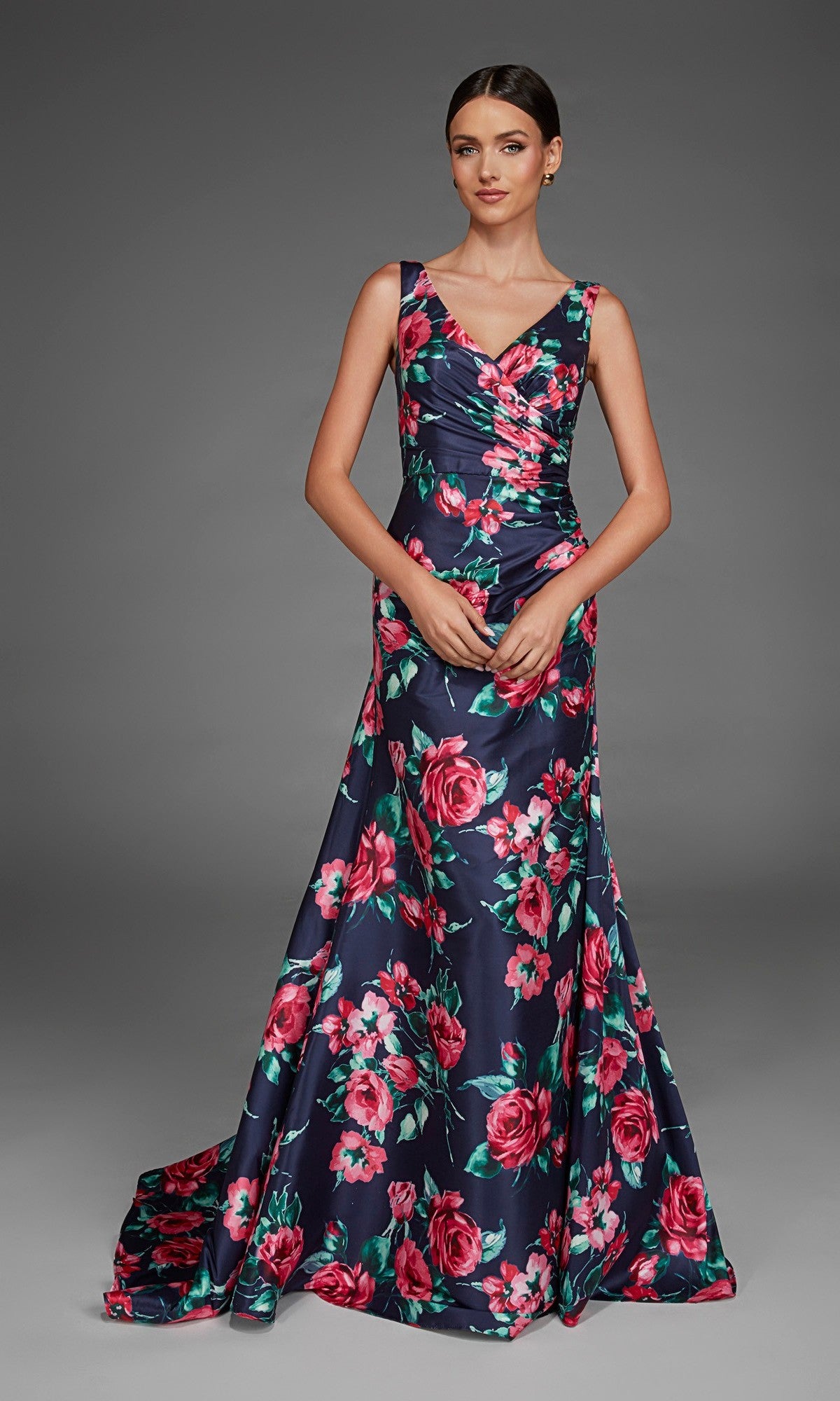 A woman wearing the Alyce 27917 long formal dress in Midnight—a floor-length, sleeveless V-neck evening gown featuring a bold red and pink floral pattern on a dark background—stands against a plain gray backdrop.