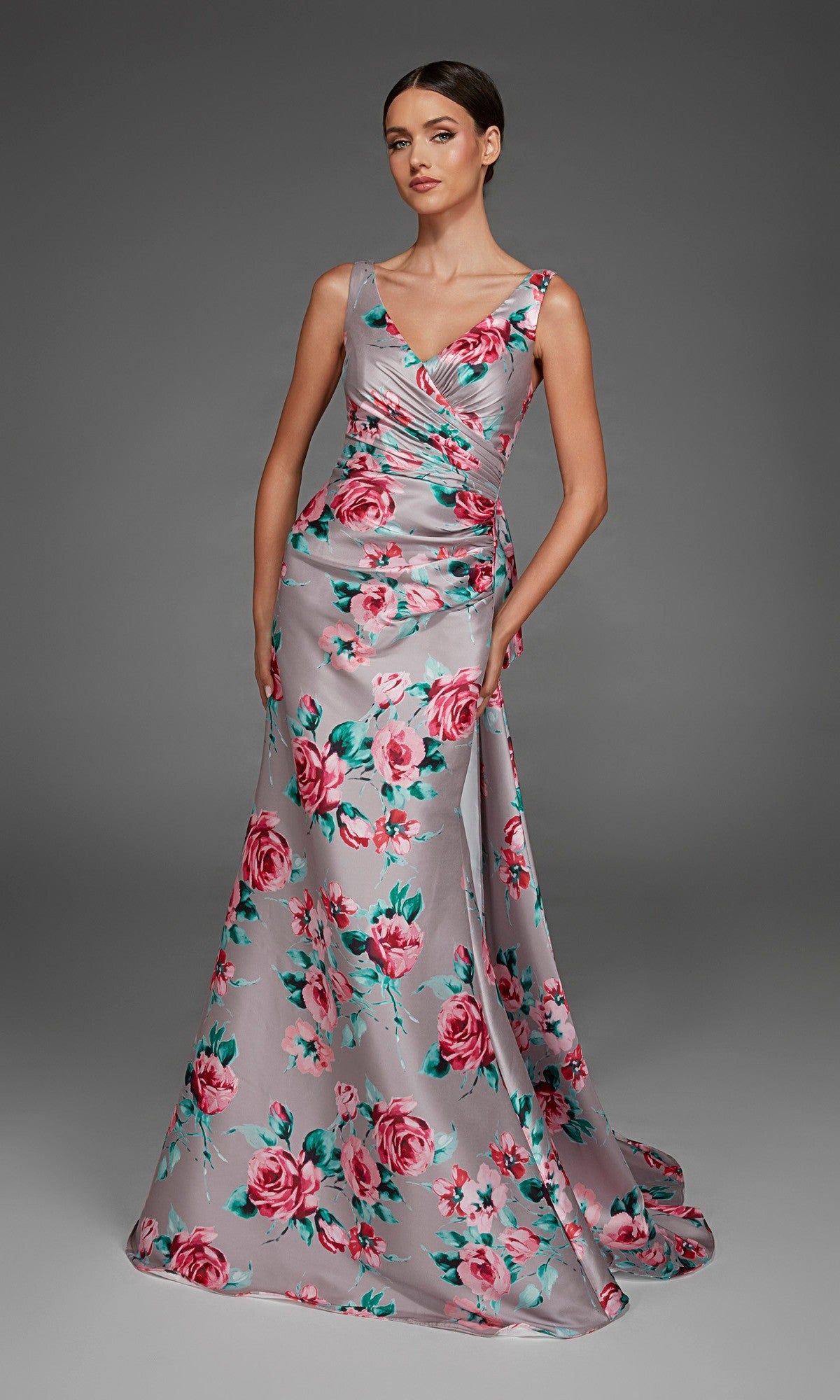 A woman wearing the Alyce 27917 long formal dress in Soft Heather, a floor-length, sleeveless silver v-neck evening gown with pink and green floral print, poses against a plain gray background.