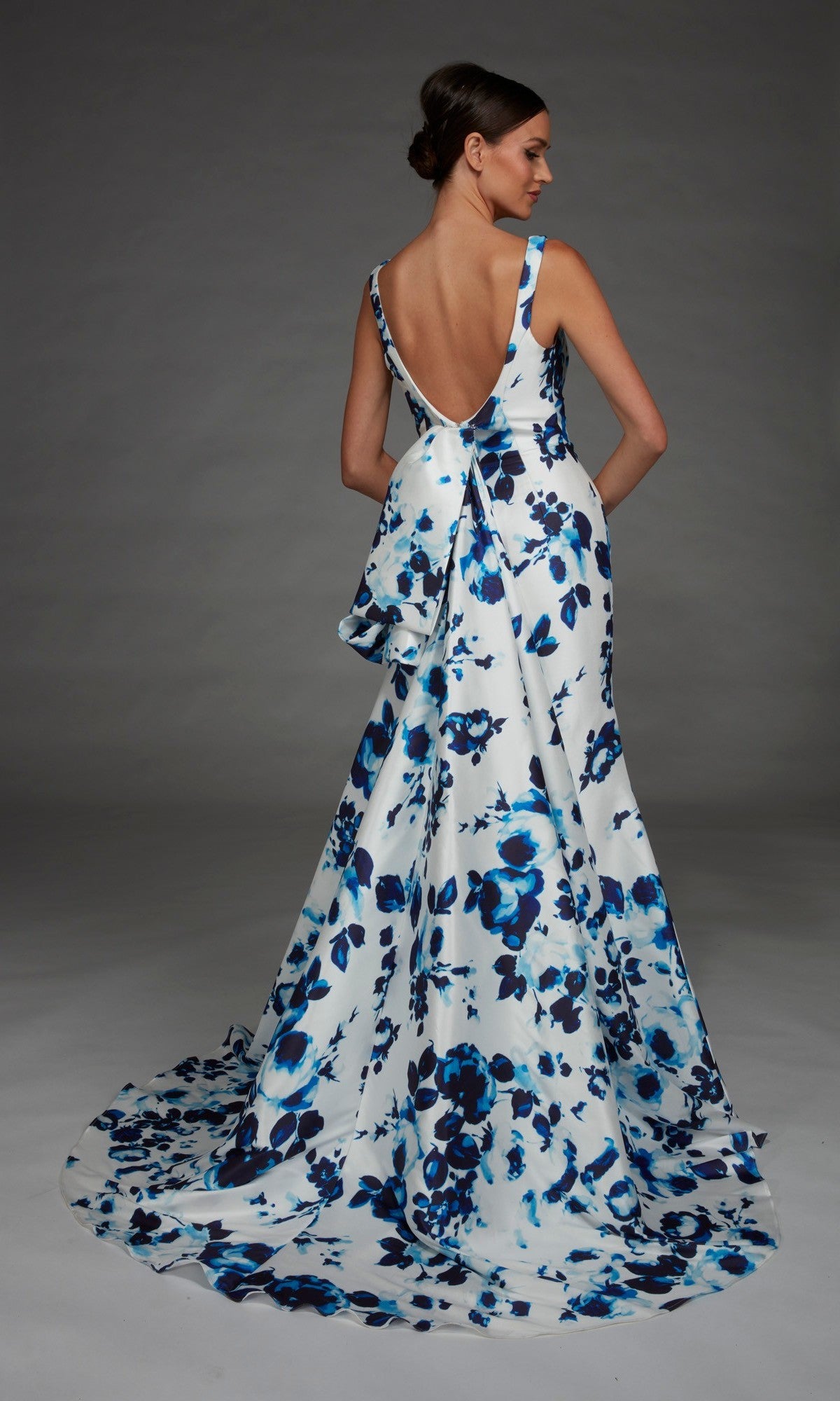 A woman wearing the Long Formal Dress: Alyce 27917—a dramatic, floor-length, backless white evening gown featuring a blue floral print and a large bow—stands against a plain gray background.