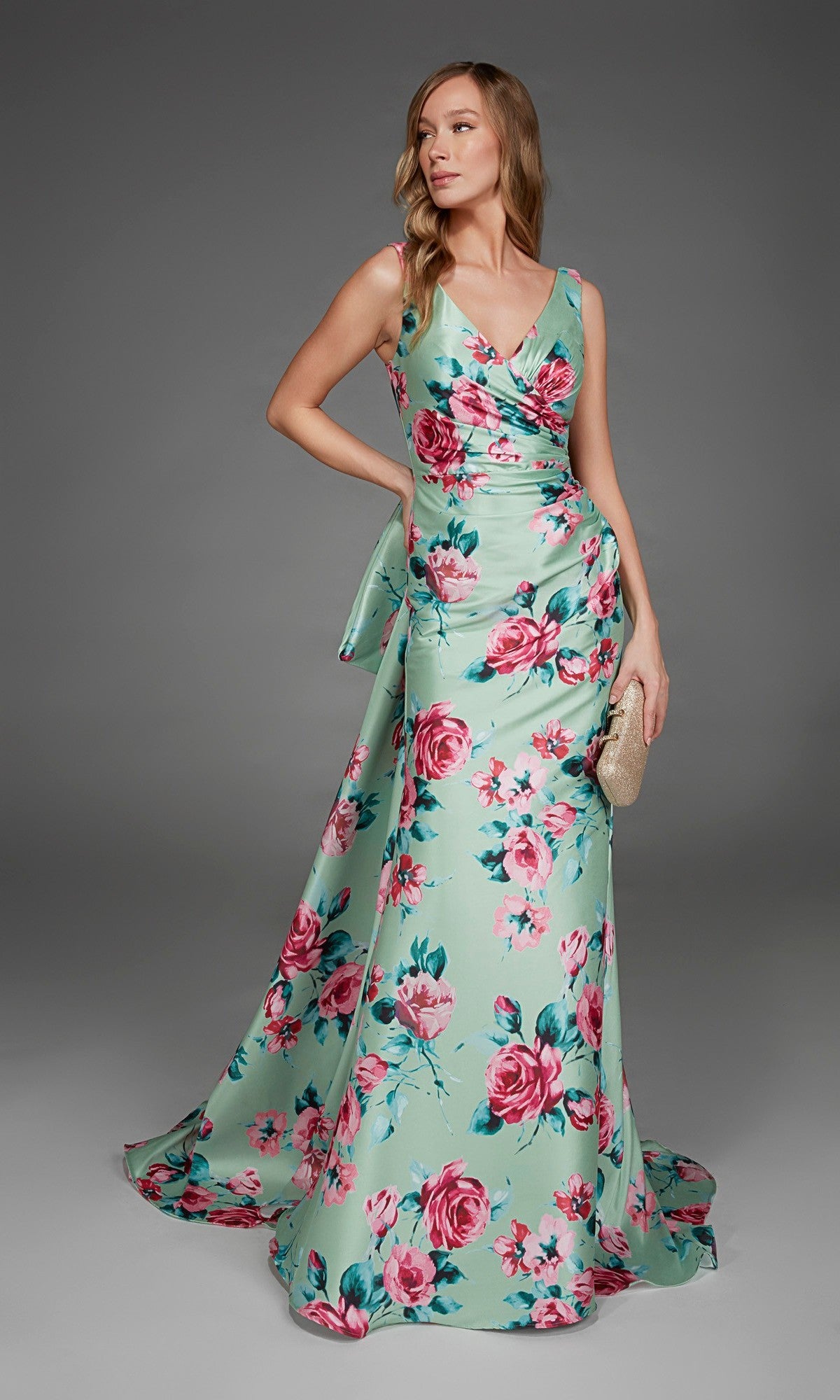 A woman poses against a plain gray background wearing the Alyce 27917 long formal dress—a sleeveless, mint green v-neck evening gown with a pink floral print—while holding a beige clutch.