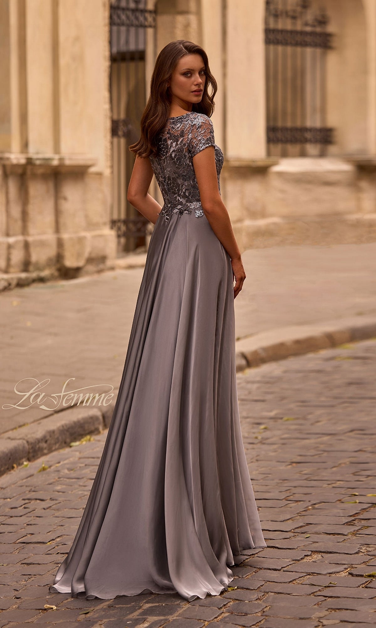 Woman wearing Long Formal Dress: La Femme 27924 with metallic lace bodice and short sleeves, standing on a cobblestone street by a stone building.