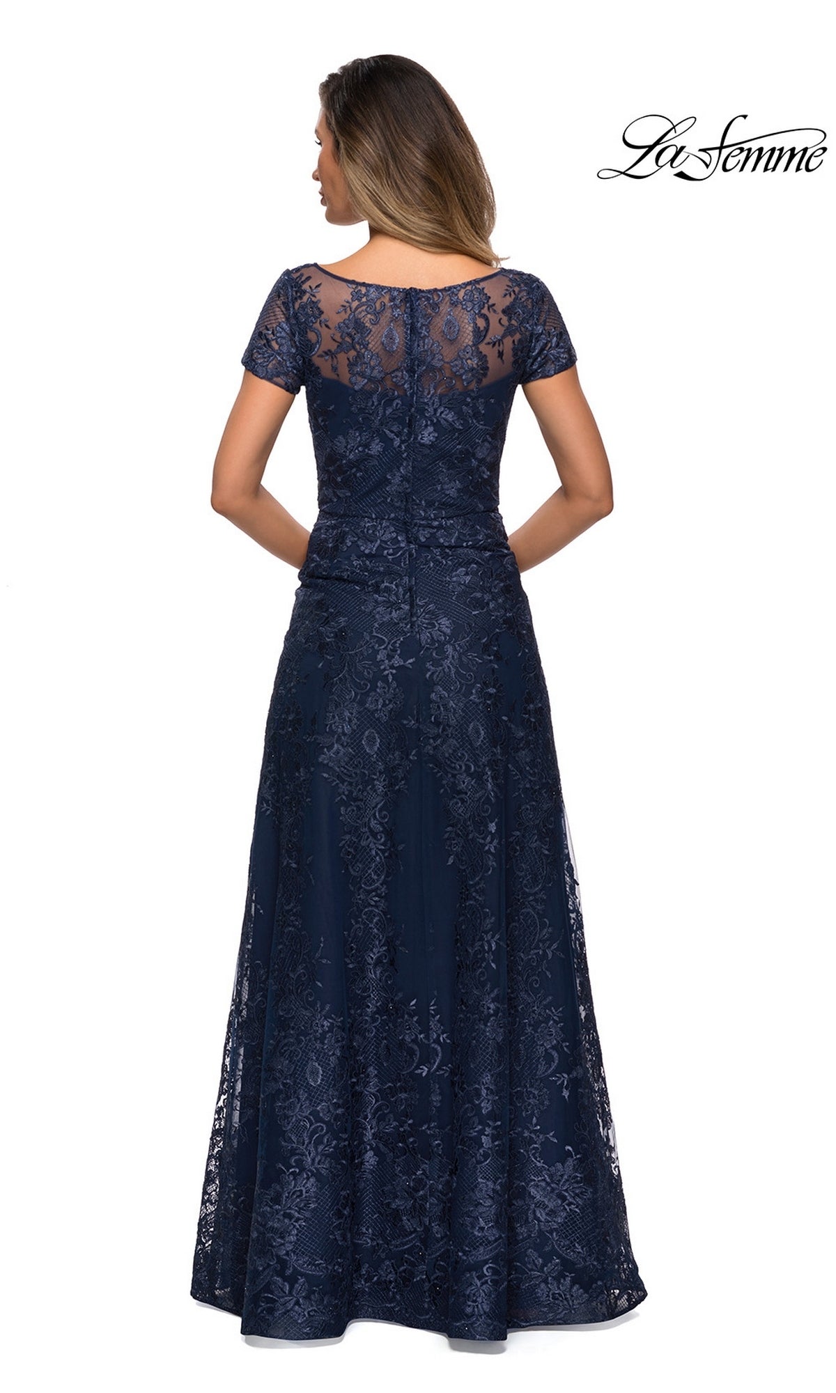 A woman stands facing away, wearing the Long Formal Dress: La Femme 27935 in navy blue with short sleeves and a sheer floral lace back.