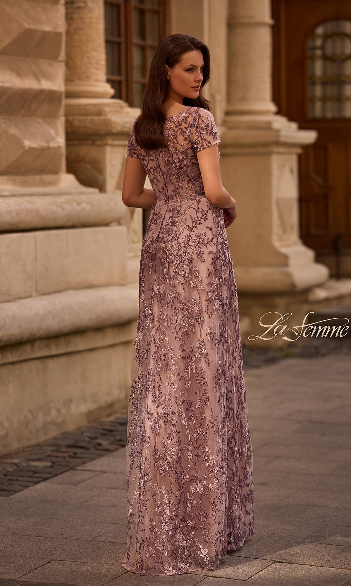 A woman wearing the Long Formal Dress: La Femme 27956 with lavender lace and floral beading stands on a cobblestone street, facing away near a stone building.