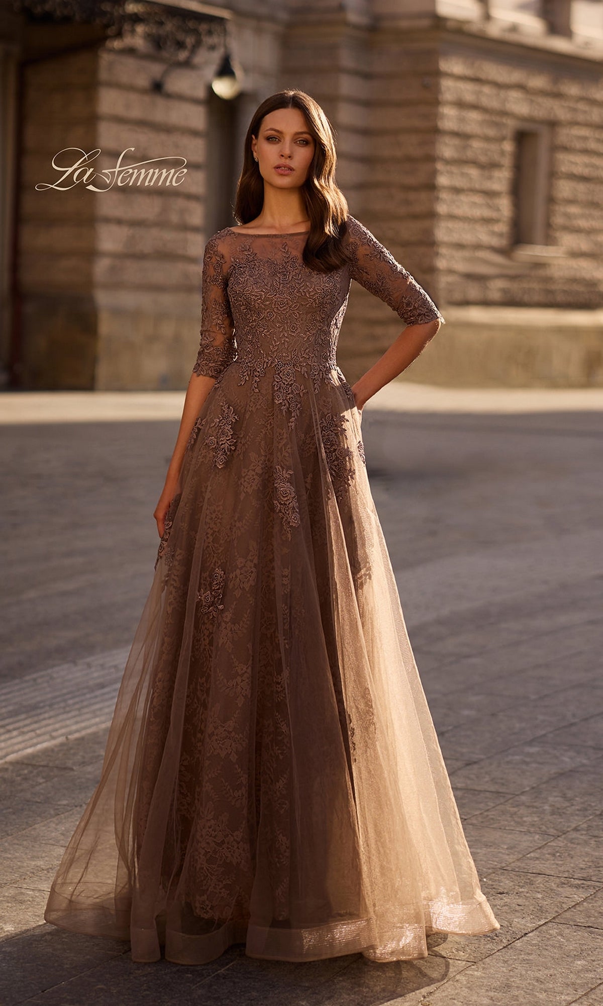 A woman stands outdoors in the Long Formal Dress: La Femme 28036 in Cocoa, featuring brown floral lace applique, three-quarter sleeves, and a flowing tulle skirt.