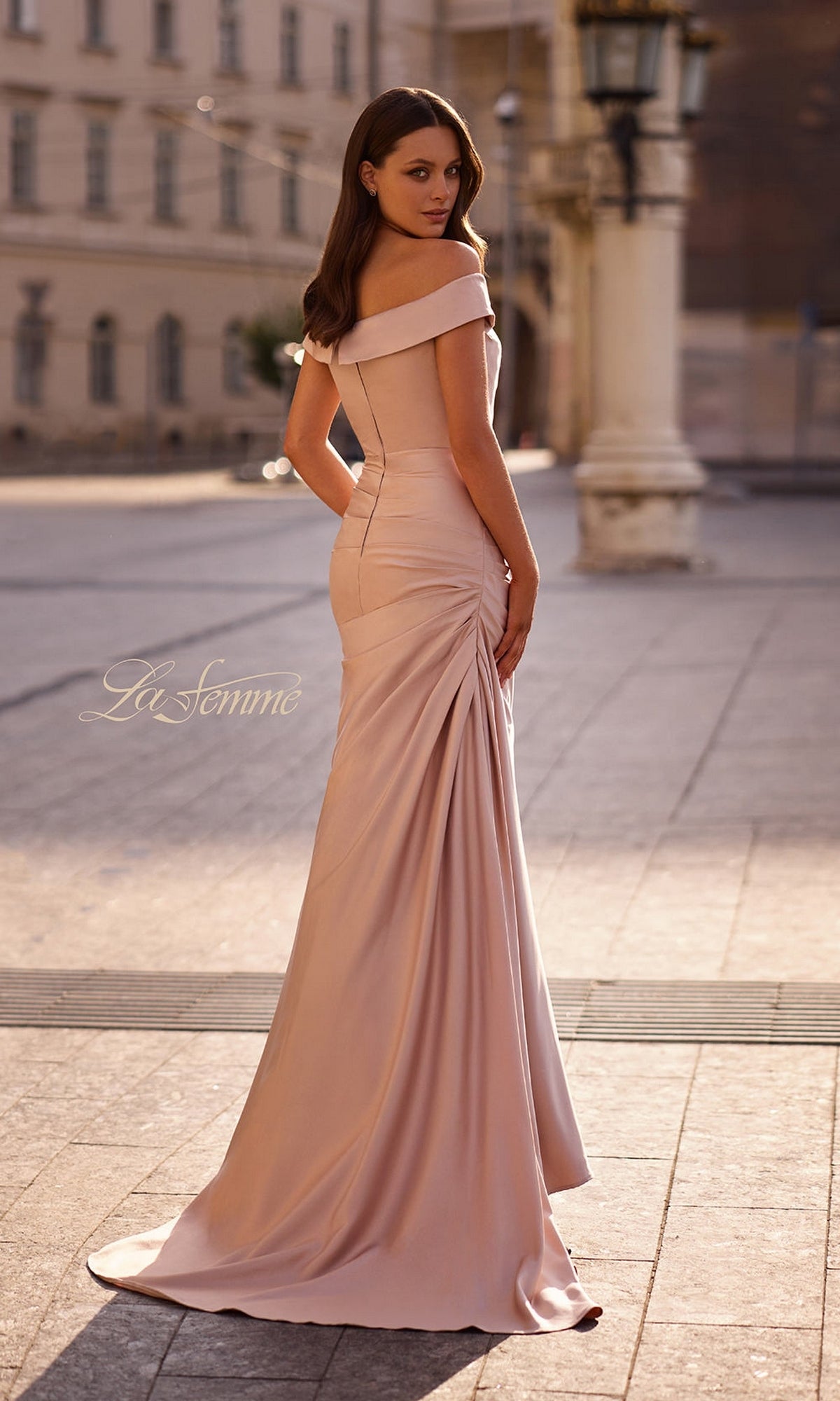 A woman wearing the La Femme 28103 long formal dress with an off-the-shoulder neckline stands on a sunlit city street, looking over her shoulder.