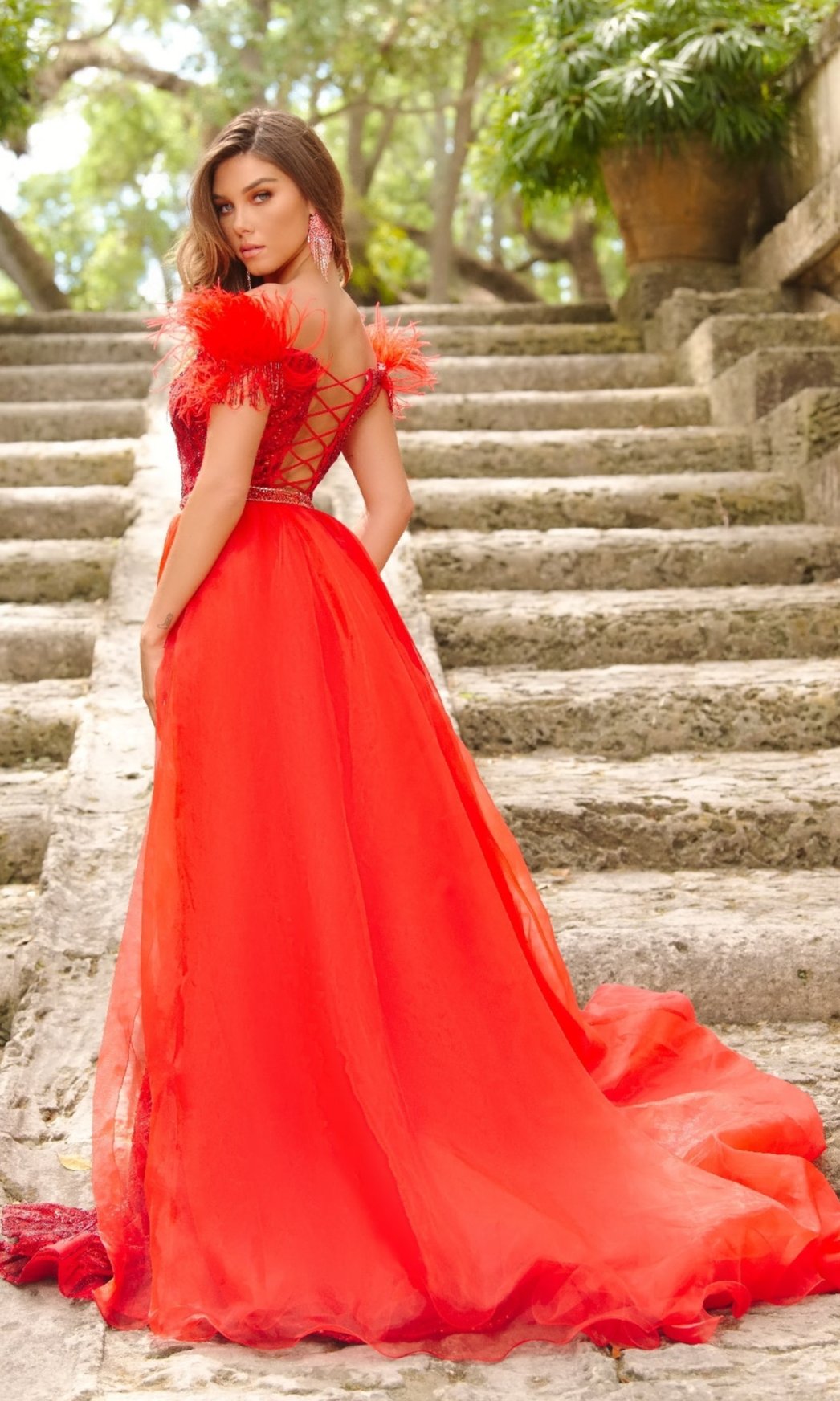 A woman stands on stone steps outdoors amid lush greenery, wearing the Long Formal Dress 28221 by Ava Presley—a bright red, off-the-shoulder mermaid gown with a lace-up back—glancing elegantly over her shoulder.