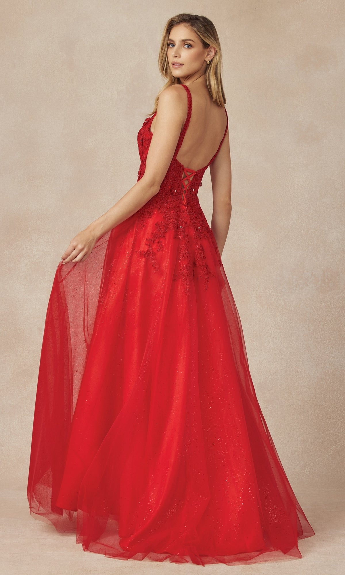 A woman wearing the Long Prom Dress 283 by Juliet—sleeveless, floor-length red A-line with a low back, sheer overlay, and lace embroidery—stands against a neutral textured background, looking over her shoulder.