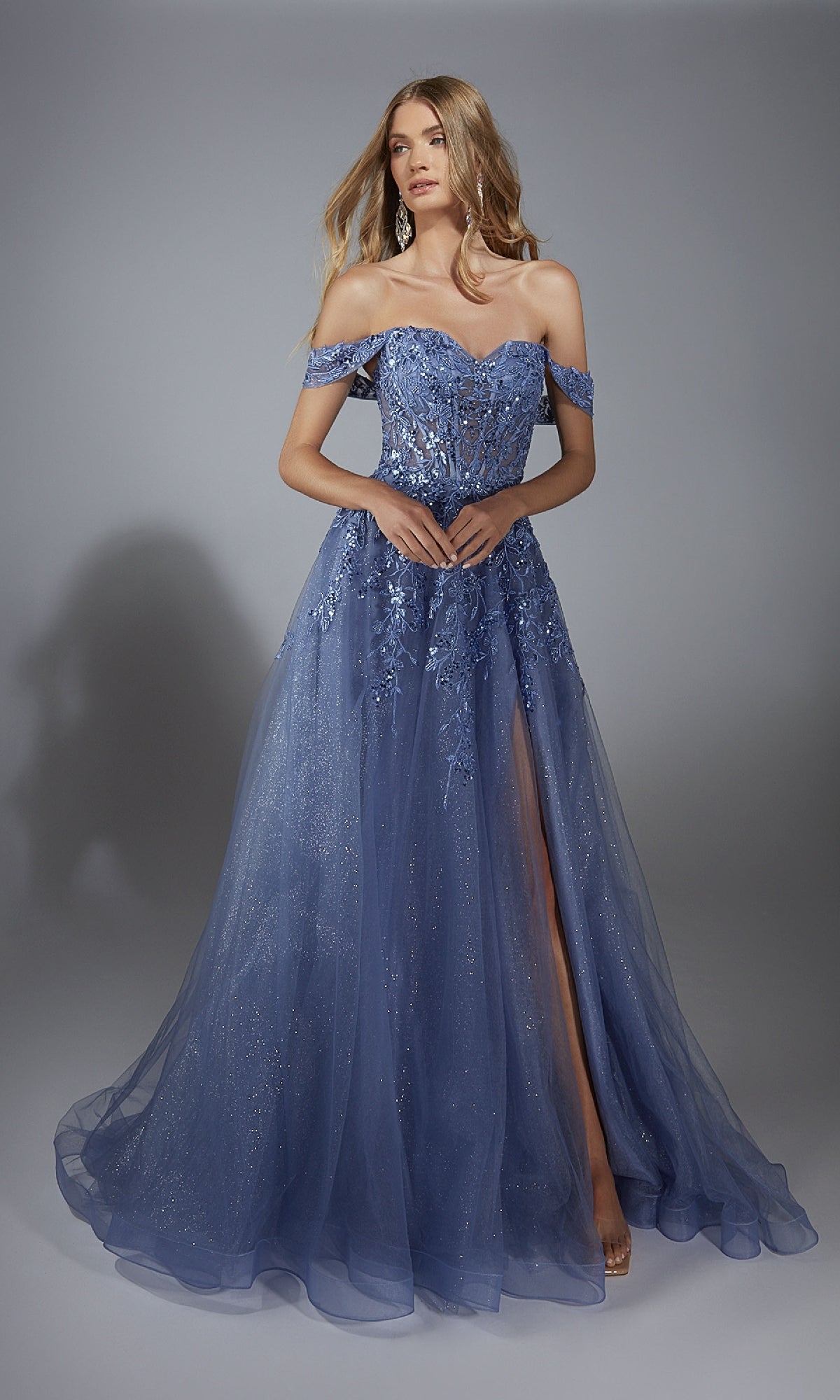 A woman stands against a plain background wearing the Aerie Couture 284 long prom dress—an off-the-shoulder, floor-length blue gown with floral embroidery, a slit, corset bodice, and an a-line skirt with tulle overlay.