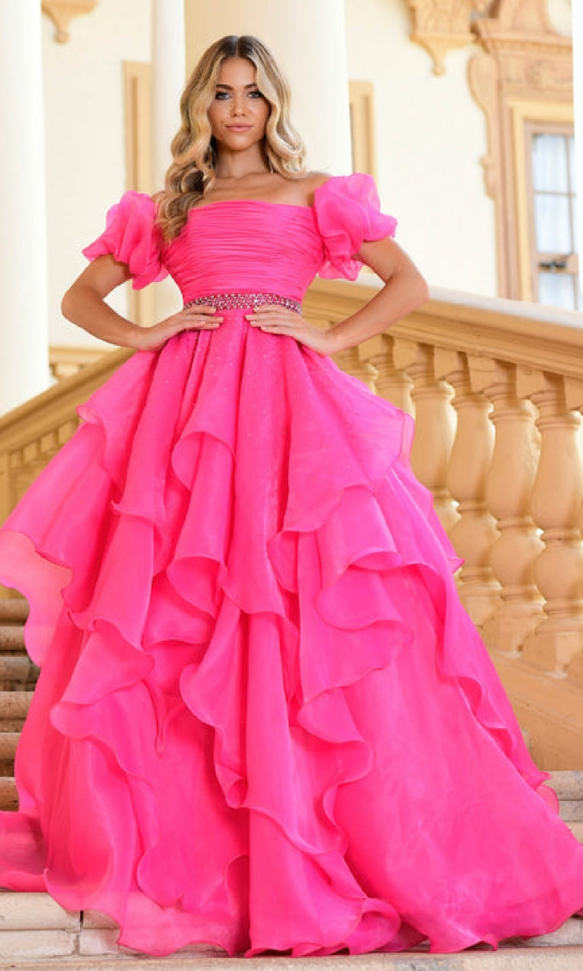 A woman stands on stairs in a bright pink Puff-Sleeve Long Prom Ball Gown (Ava Presley 28571) with ruffles and a jeweled belt, styled for elegance, with a classical building as the backdrop.