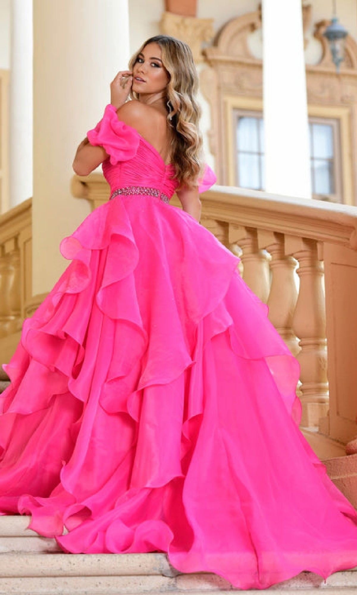 A woman in a vibrant pink Ava Presley 28571 puff-sleeve long prom ball gown stands on a staircase, framed by ornate columns and a grand building façade.