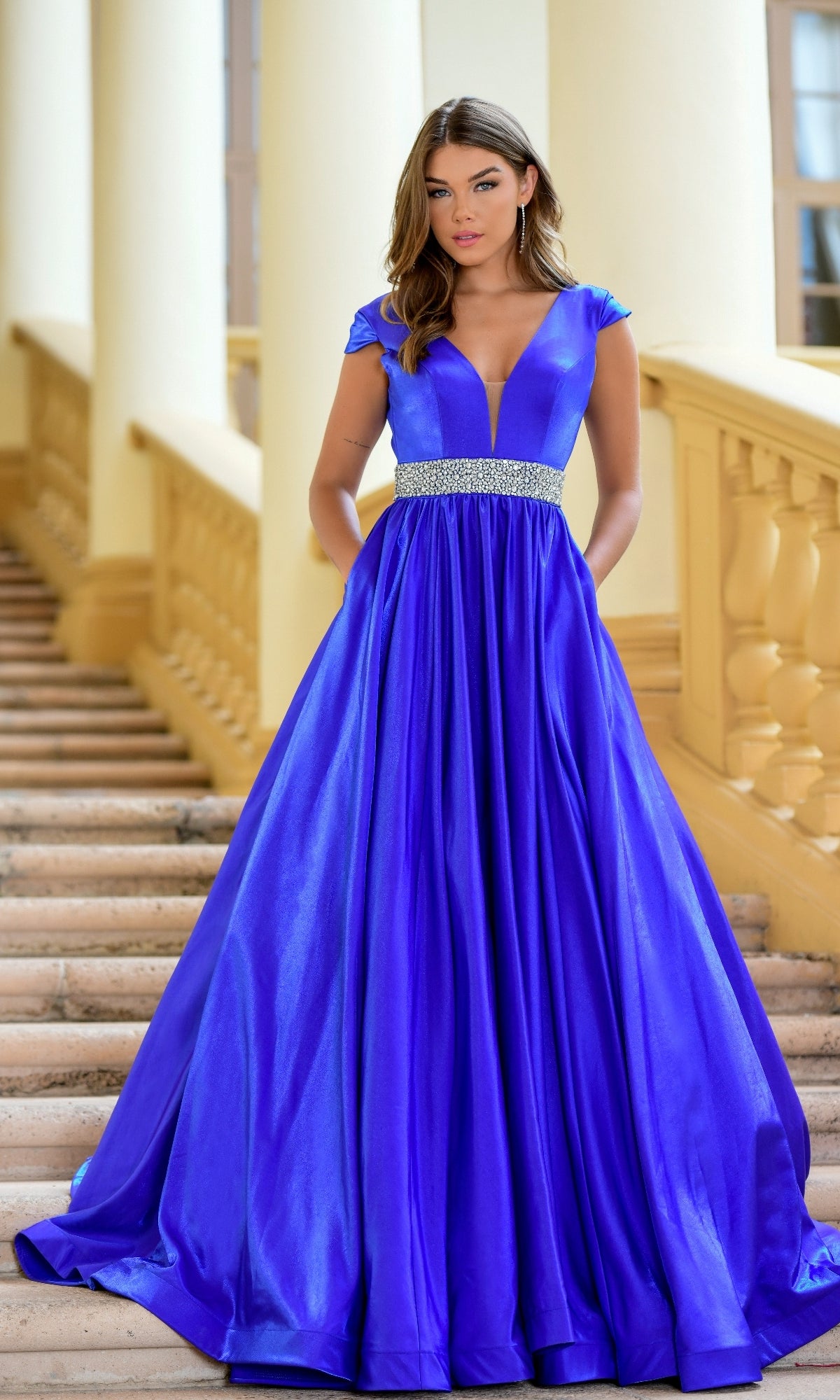 A woman stands on stairs with a cream railing and columns, wearing the Long Formal Dress 28580 by Ava Presley—a royal blue, floor-length A-line gown featuring a deep V-neckline and jeweled waistband.