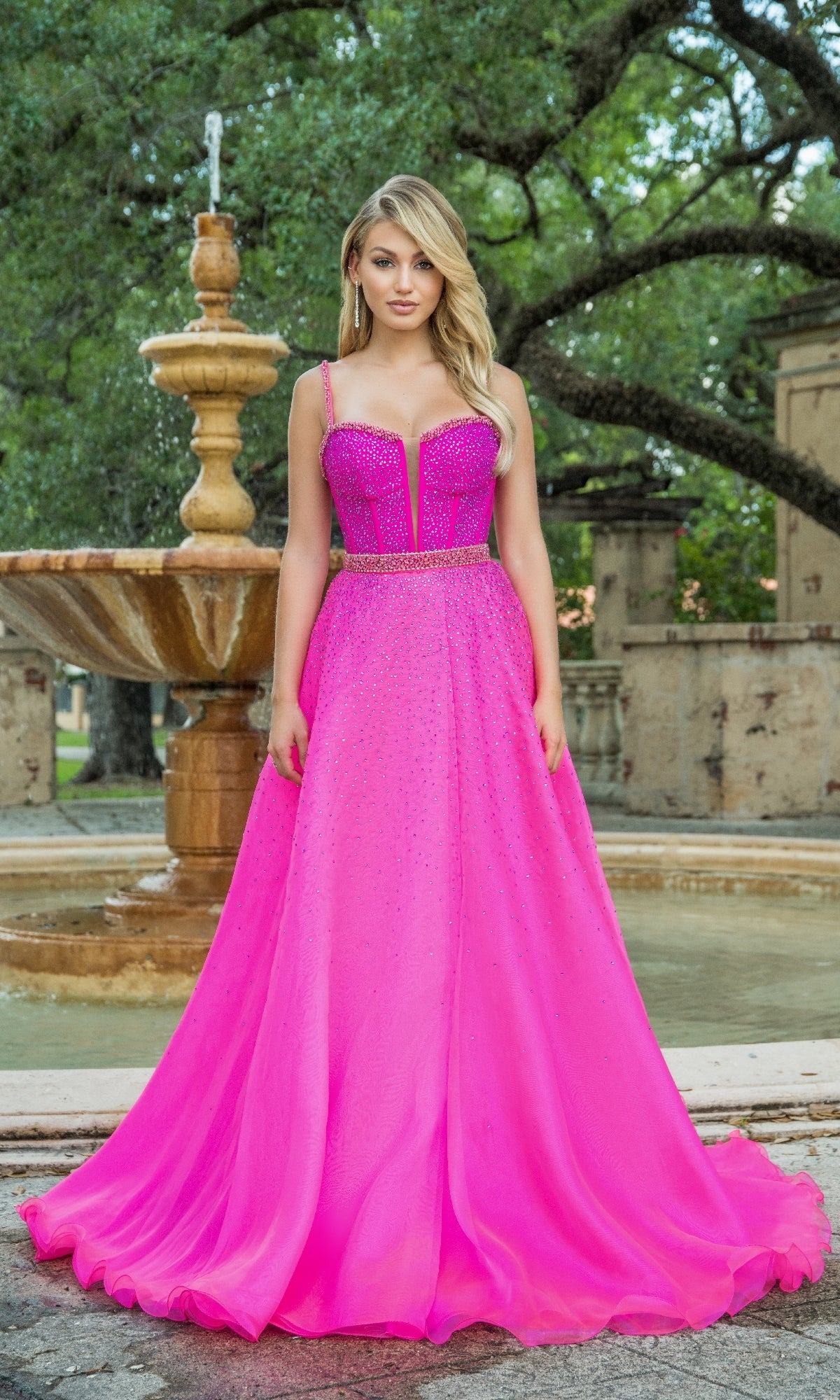 A woman stands outdoors by a fountain, wearing the Ava Presley Long Formal Dress 28581—a bright pink gown with beaded details, a fitted corset bodice, and a flowing floor-length skirt.