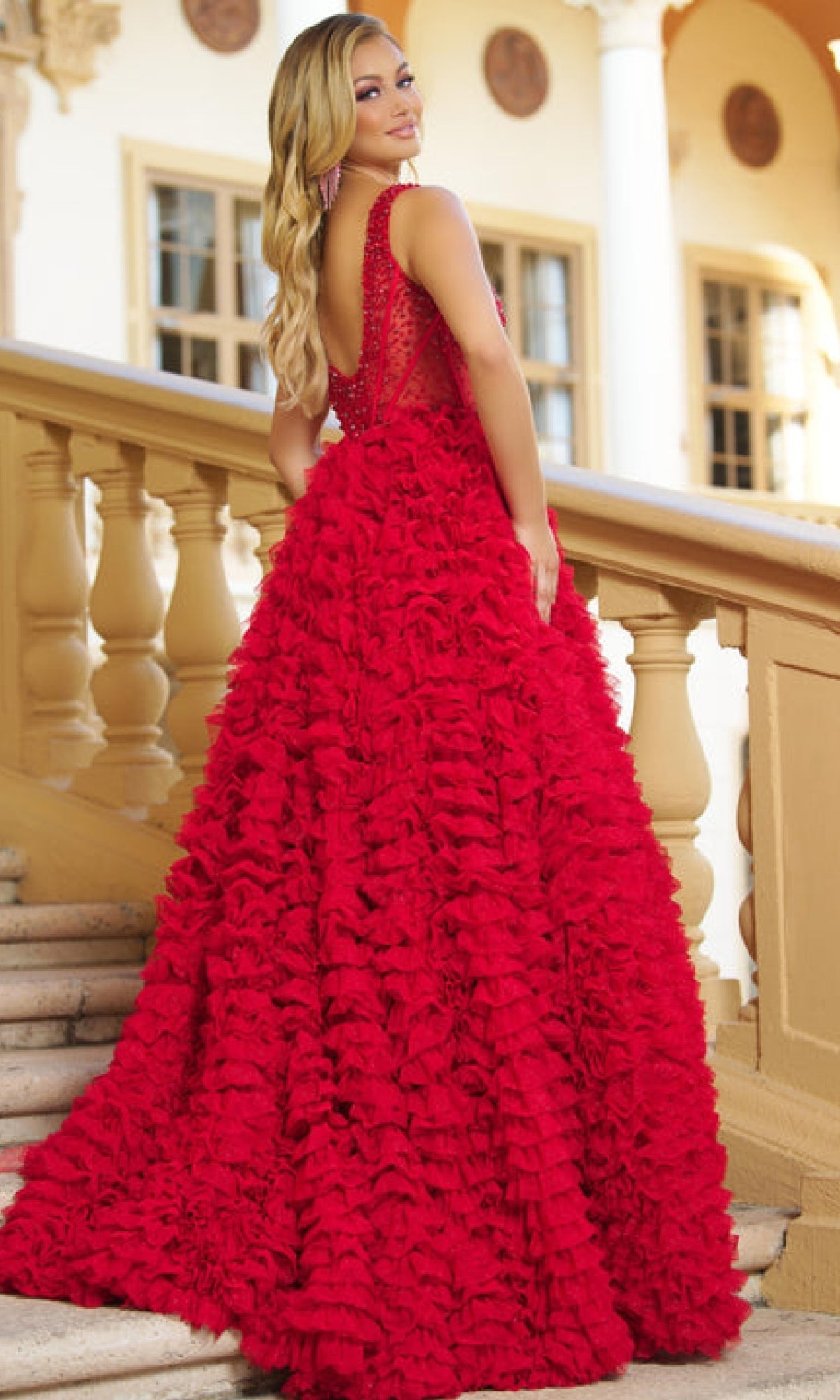 On a staircase, a woman looks over her shoulder in elegant confidence, wearing the Ava Presley 28584 long ruffled prom ball gown with a sheer back.