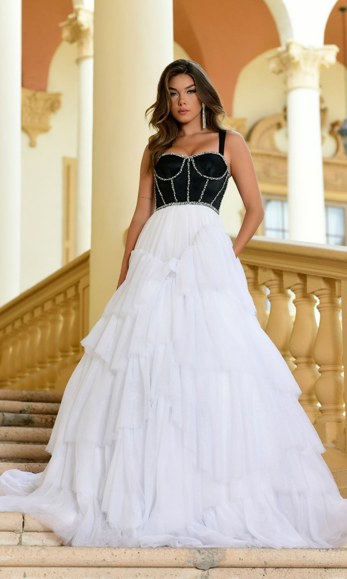 A woman stands on stairs wearing the Long Formal Dress 28592 by Ava Presley, featuring a black corset bodice and a voluminous white tiered tulle skirt, with columns and arches in the background.