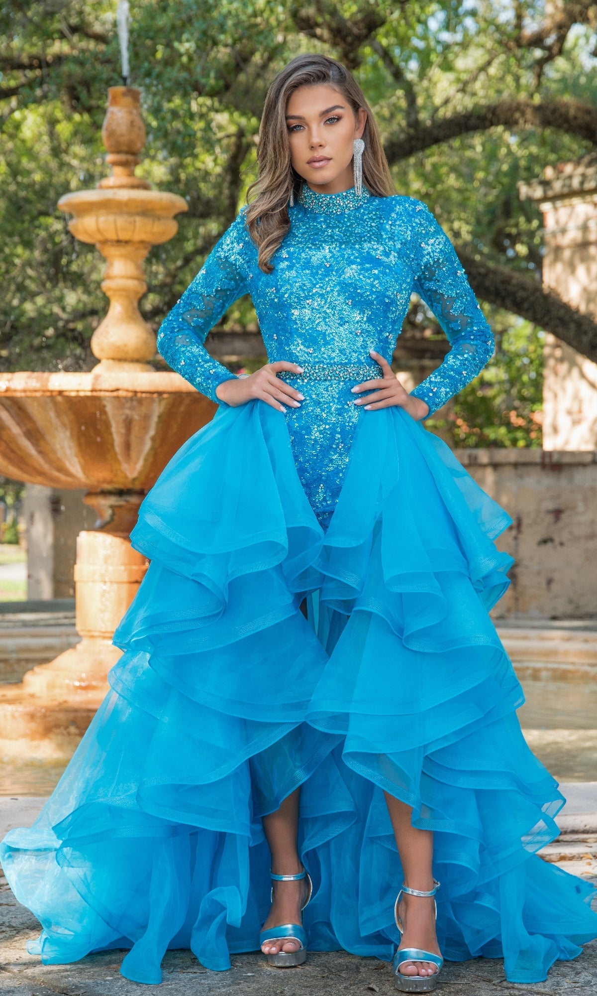 A woman stands outdoors before a fountain, wearing the Ava Presley 28595 Ruffle Short Sequin Romper and silver high-heeled sandals.